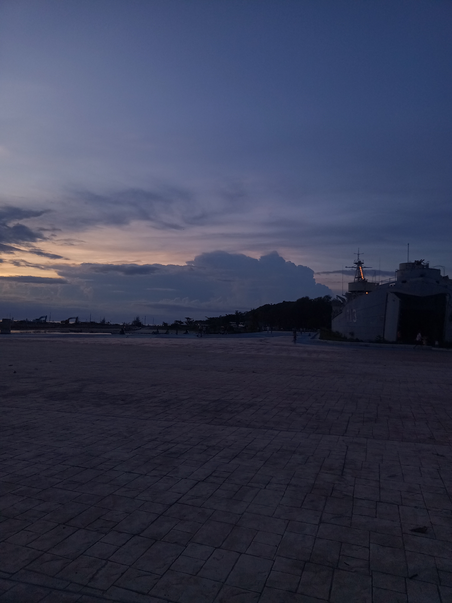 This is a vertical, medium-shot photograph taken at dusk. The sky is a deep, dusky blue with dramatic, heavy clouds stretching across the horizon. A pale orange glow peeks through the clouds on the left, indicating the last rays of sunset.
In the foreground, a paved area made of rectangular stone tiles occupies the lower half of the frame. The texture of the tiles is clearly visible, and they appear to be a dark gray color.
In the middle ground, to the right, a large, gray ship is docked. It has a prominent superstructure with antennas and lights. The number "113" is visible on the side of the ship. The ship is imposing and appears to be a naval vessel. To the left of the ship, a line of trees forms a dark silhouette against the twilight sky. Further to the left, there are hints of construction equipment, possibly excavators, and other structures along the shoreline.
A few small figures of people are scattered across the paved area and near the ship. They are silhouetted against the fading light, and their activity is not clearly discernible, but they appear to be walking or standing.
The overall mood of the image is serene and slightly melancholic, characteristic of twilight. The location cues, such as the ship and the tropical trees, along with the provided context of Koh Phangan, Thailand, suggest a coastal or port setting. The weather appears calm, with the clouds being the most dynamic element in the scene.