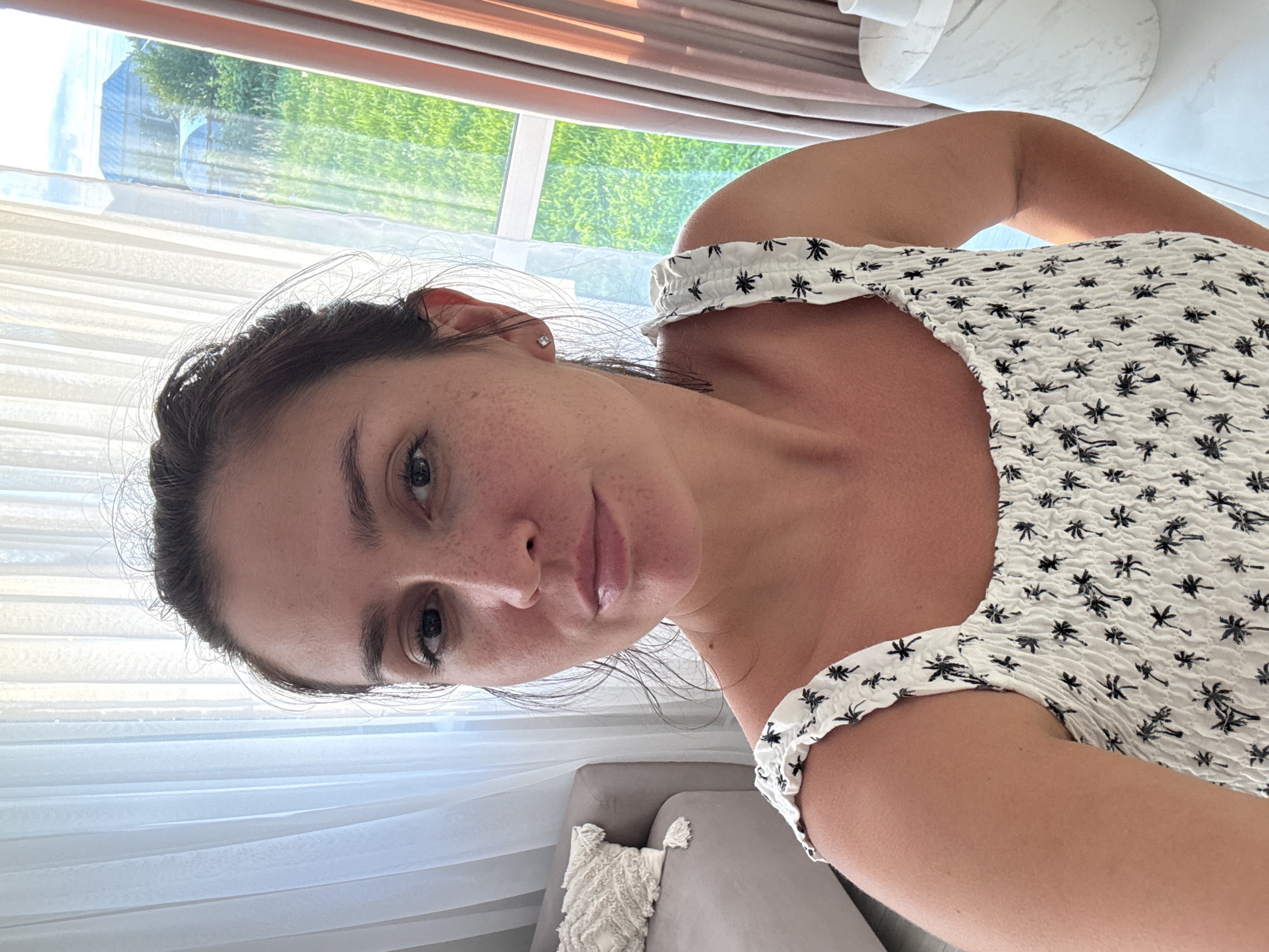 The image is a medium shot selfie of a woman in her late 20s or early 30s with her hair pulled up. She is wearing a white smocked dress with a black palm tree pattern. The woman has light skin with freckles and dark hair. She is looking directly at the camera with a neutral expression. Behind her, to the left, is a gray sofa with a decorative pillow. To the right, a window with sheer white curtains on the left and pinkish-brown drapes on the right reveals a green, grassy outdoor area with some structures visible in the distance, possibly in the city of Nizhnyayarkeyevo, Russia. The lighting suggests it is daytime, with sunlight filtering through the window.