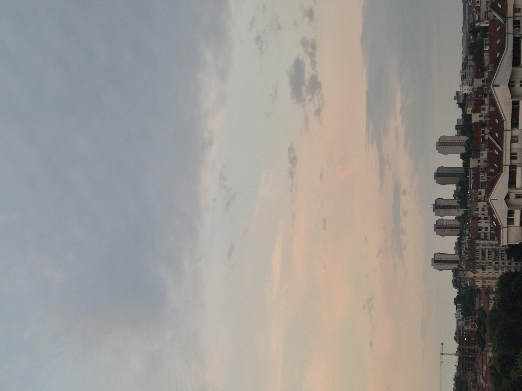 This is a medium-angle, landscape-oriented outdoor photograph of a city skyline during twilight. The sky occupies the top two-thirds of the frame and is filled with clouds, transitioning from a pale grey at the top to soft pinks and oranges closer to the horizon. Below the sky, the city is visible, characterized by a dense collection of buildings. Several taller, modern apartment buildings with a distinctive stepped facade are prominent in the mid-ground, arranged in a cluster. In the foreground and to the left, there are lower-rise residential buildings with red-tiled roofs and visible windows. A construction crane is also visible on the left side of the frame, indicating ongoing development. Trees are interspersed throughout the cityscape, particularly in the foreground and between buildings. The overall lighting suggests either sunrise or sunset, with a diffused, soft light illuminating the scene.