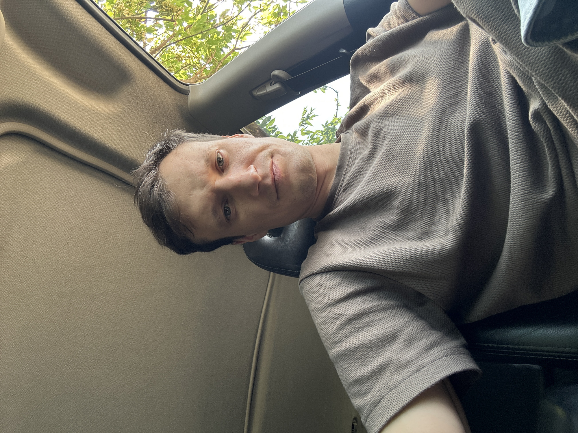A medium shot captures a man from the chest up, seated in the driver's seat of a car. He is looking directly at the camera with a neutral expression. Sunlight streams in from the right, illuminating his face and the textured fabric of his taupe-colored shirt. The car's interior is visible, with the headliner and a portion of the sunroof above him. Outside the car, green foliage is visible, suggesting a location with trees. The image was taken in Makiv, Ukraine.