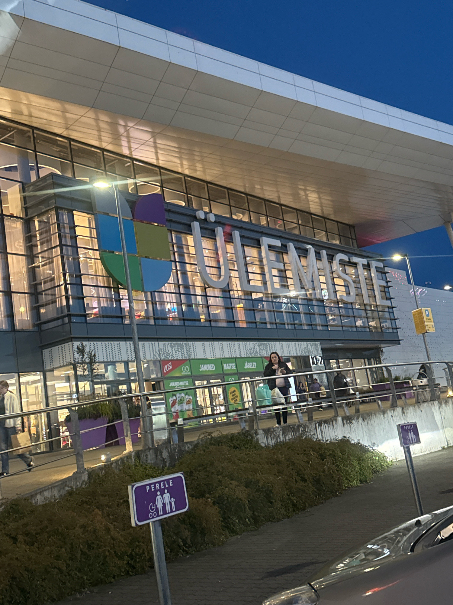 The image shows the Ülemiste Center in Tallinn, Estonia, at dusk. The building is a large, modern shopping mall with a facade of glass and white panels. The name of the center, "ÜLEMISTE," is displayed prominently in large, illuminated letters across the upper part of the building. Below the main signage, there is a lower level with shop entrances and windows, some of which are lit.
In the foreground, to the right, a purple sign with a white silhouette of a family and a wheelchair indicates a "PERELE" (family) parking area. A dark-colored car is partially visible in the bottom right corner, reflecting the ambient light.
On a raised walkway in front of the shopping center, several people are visible. One woman, wearing a dark jacket and carrying a light-colored bag, stands near the entrance. To her left, a person in a light-colored jacket and dark pants is walking away from the camera, carrying a shopping bag. Other individuals are seen in the background, either walking or standing on the walkway.
The area in front of the walkway is landscaped with bushes and large purple planters. The sky above is a deep blue, indicating it is either early evening or late dusk. Streetlights and lights from within the building illuminate the scene. The overall impression is that of a busy commercial area at the end of the day.