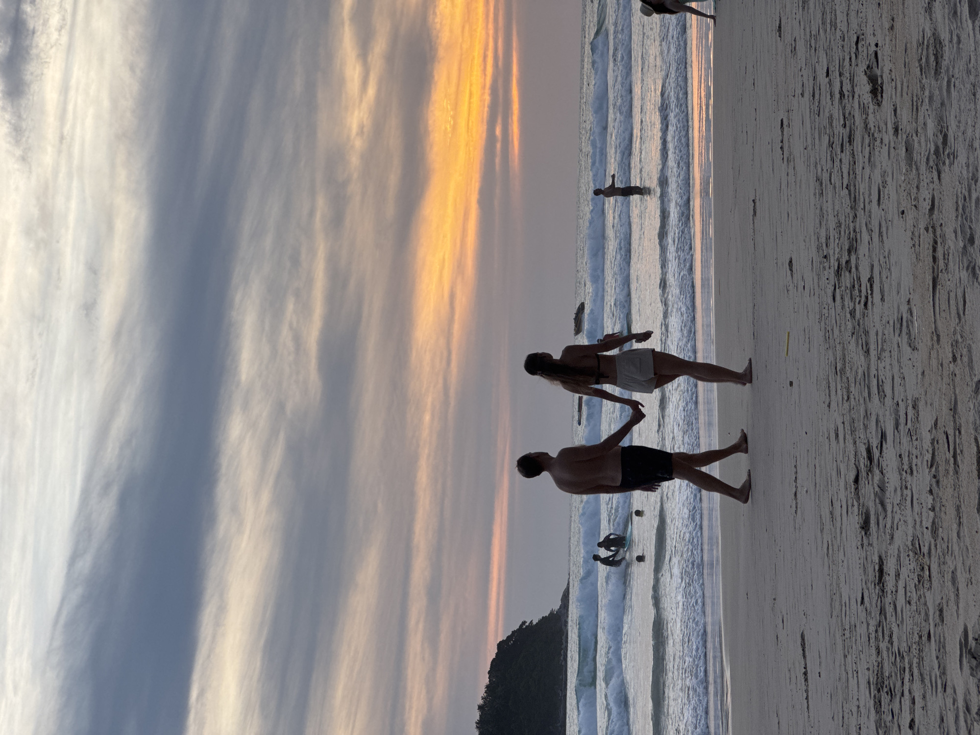 This image captures a serene sunset scene on Karon Beach, Thailand. The sky is ablaze with hues of orange and pink, streaked with soft gray clouds. Gentle waves lap against the shore, where a sandy beach stretches into the foreground. In the mid-ground, a couple walks hand-in-hand along the water's edge, their silhouettes defined against the vibrant sky. Further out, several individuals are visible in the ocean; some are wading, while others appear to be enjoying the water. A lush, green headland juts out into the sea on the left side of the frame, adding a natural contrast to the expansive sky and ocean. The overall atmosphere is one of tranquility and natural beauty.