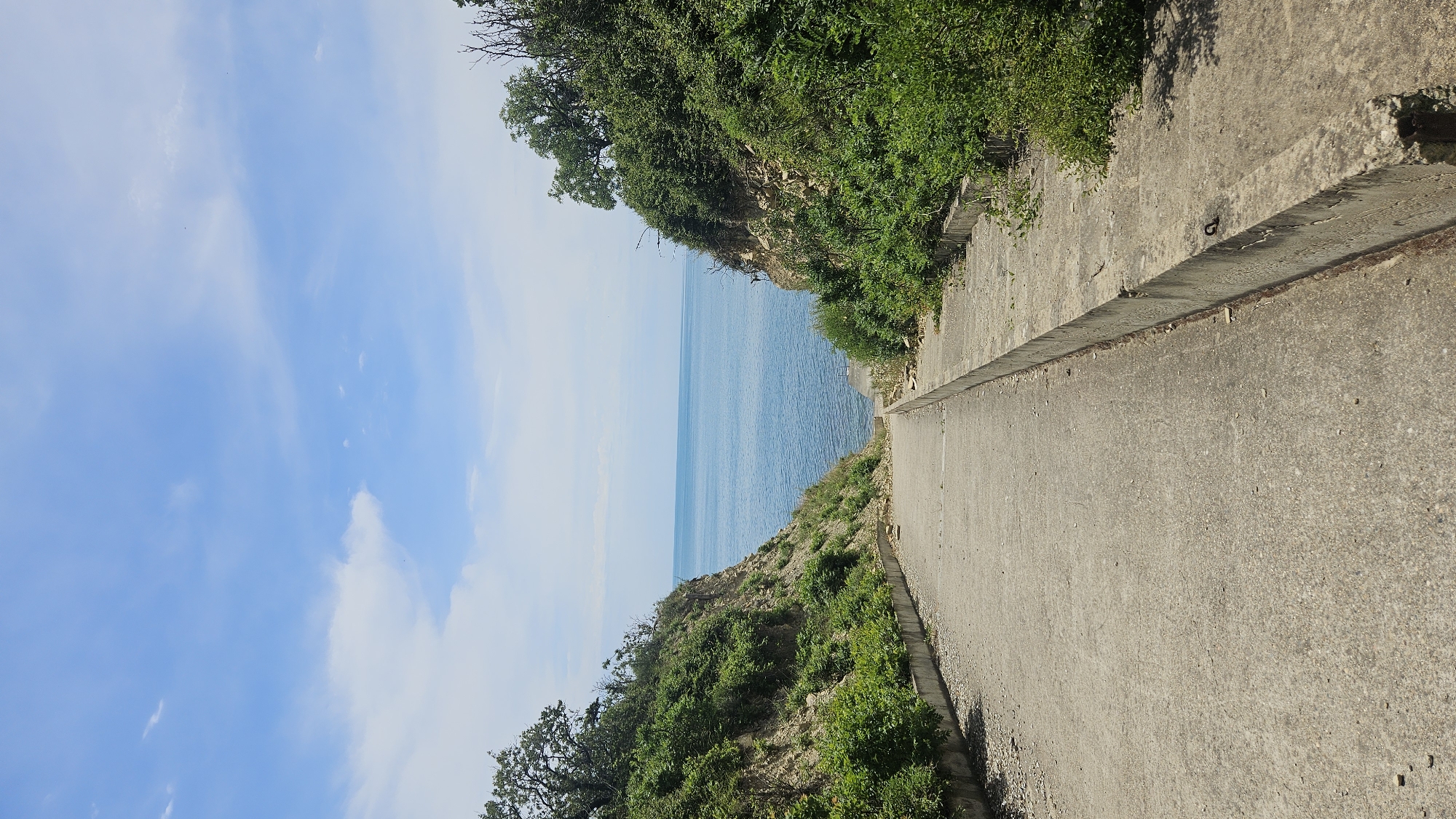 The image shows a concrete pathway leading downhill towards the Black Sea. The pathway is flanked by green, bushy vegetation on either side, with rocky cliffs visible in some areas. The sky is a clear blue with a few scattered white clouds. The sea is calm and a deep blue color, meeting the horizon in the distance. This scene is located in Dyurso, Russia.