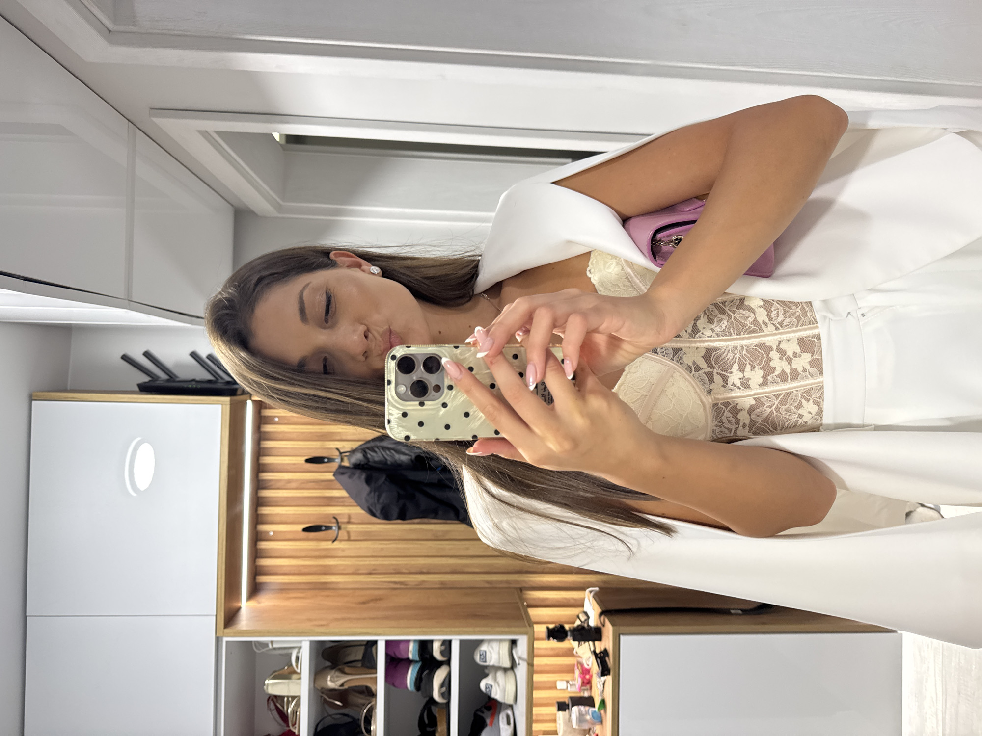 A woman takes a selfie in an interior, likely in Burgas, Bulgaria, considering the context. She holds a smartphone with a patterned case. The woman wears a white outfit with a lace bodice. She's holding a small pink purse. Behind her is a modern closet unit with white doors, wooden slats with hooks, and shoe storage. A black coat hangs on the hooks. Various pairs of shoes are visible in the shoe storage.