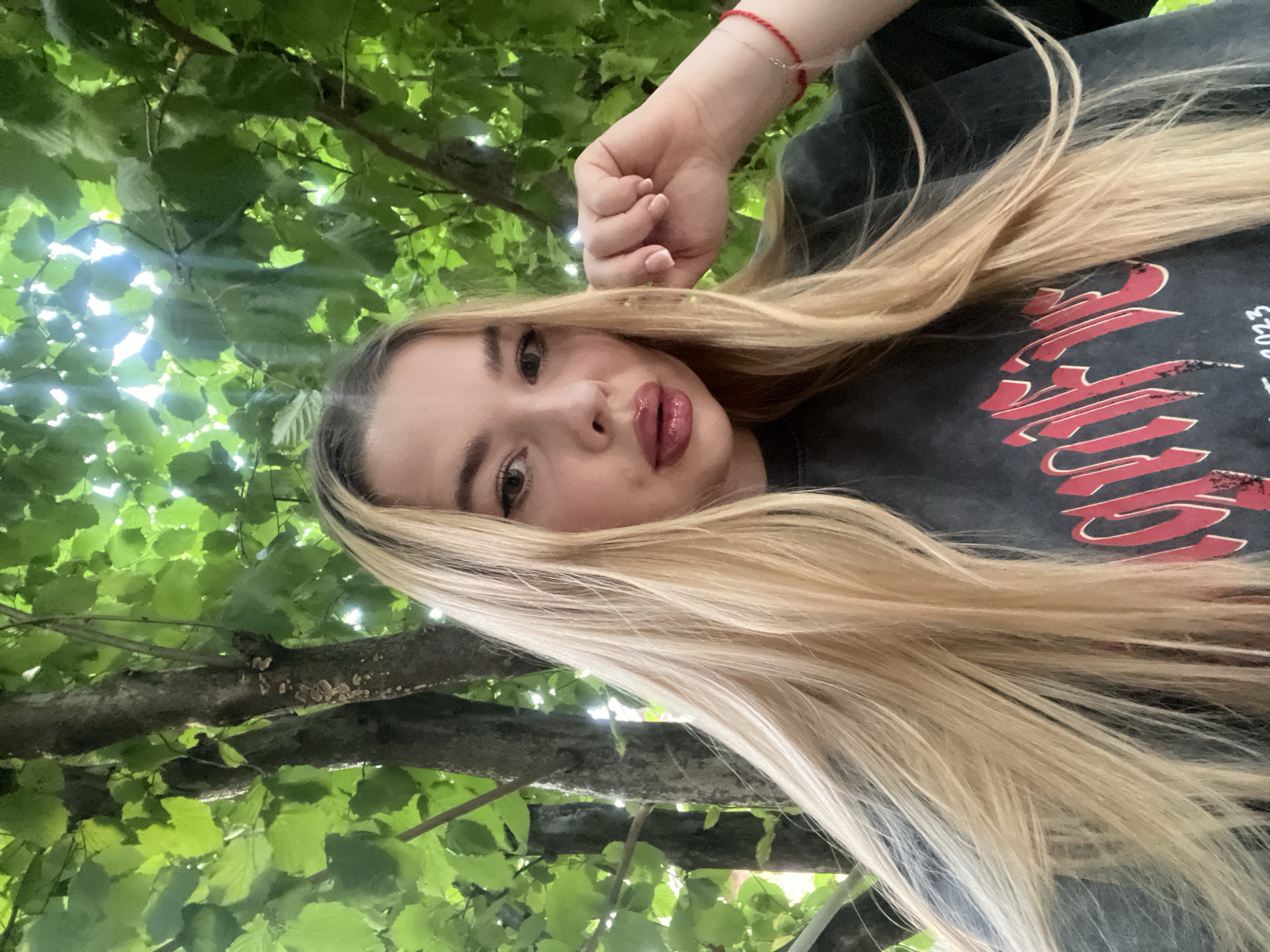 A young woman with long blonde hair is posing for a selfie outdoors. She is wearing a dark grey t-shirt with red lettering. Her right hand is raised to her face, with her fingers touching her cheek. She is wearing a red string bracelet on her left wrist. The background is filled with lush green foliage and tree trunks, suggesting a park or garden setting. There is no identifiable information to place this image in Tikhoretsk, Russia.