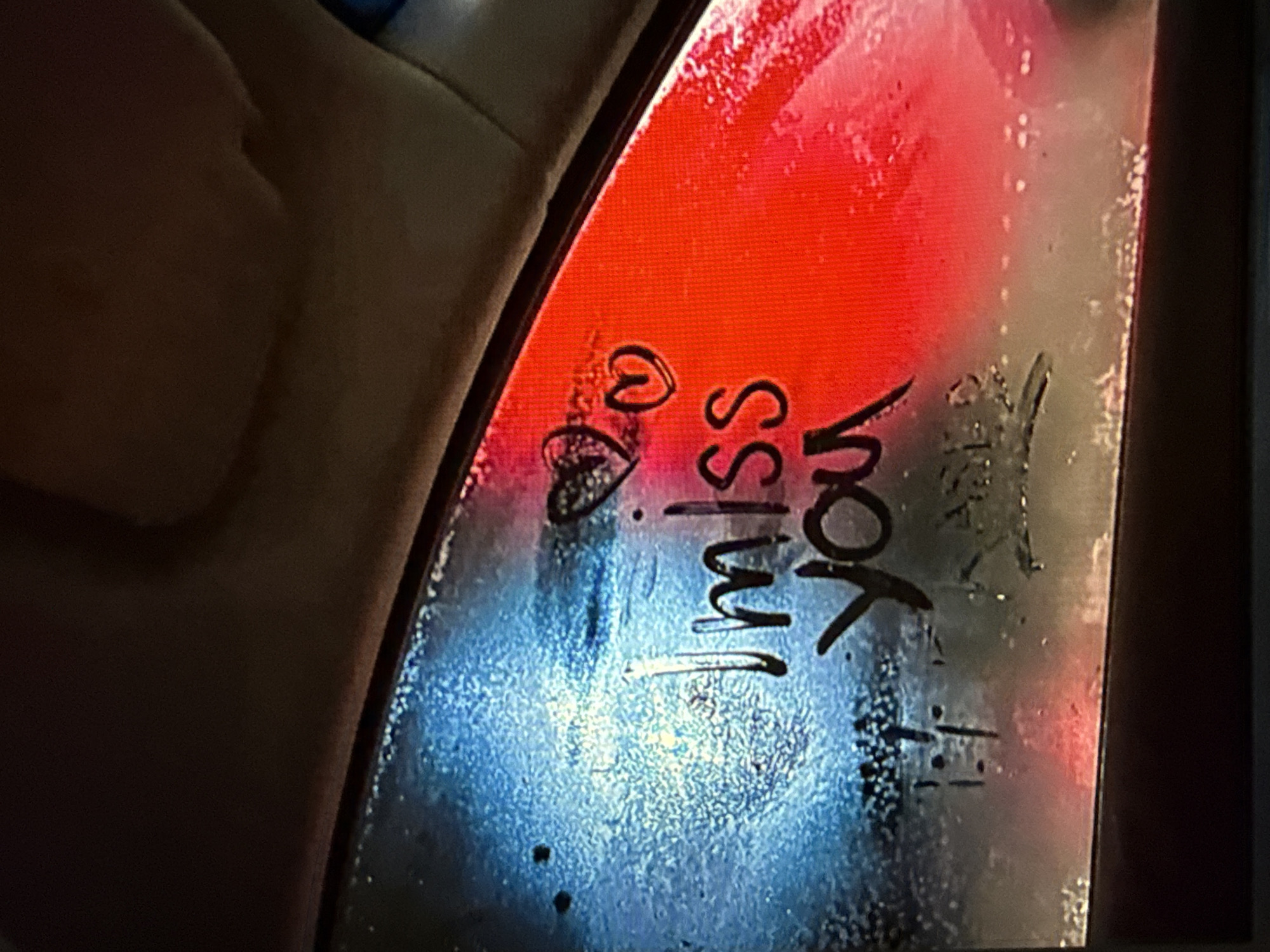 The image presents an interior perspective of a vehicle's window, which is covered with condensation. Wiped into this condensation is the handwritten message "I miss you," featuring two hearts above the text, one solid and one outlined. Faint, indecipherable marks are visible below the main text.
The exterior scene, viewed through the window, is illuminated by distinct artificial light sources. The left side of the window reflects a strong blue light, while the right side is dominated by a vibrant red light, creating a sharp color division across the glass. This suggests the vehicle is stationary amidst external lighting, possibly from traffic signals, vehicle lights, or illuminated signs in an urban setting. The interior environment appears dark, with the visible upper edge showing a light-colored headliner and the lower edge a darker window frame or door panel.
There are no individuals visible in the image, nor are there any identifiable landmarks or features to determine a specific geographical location, including the city of Kardzhali, Bulgaria.