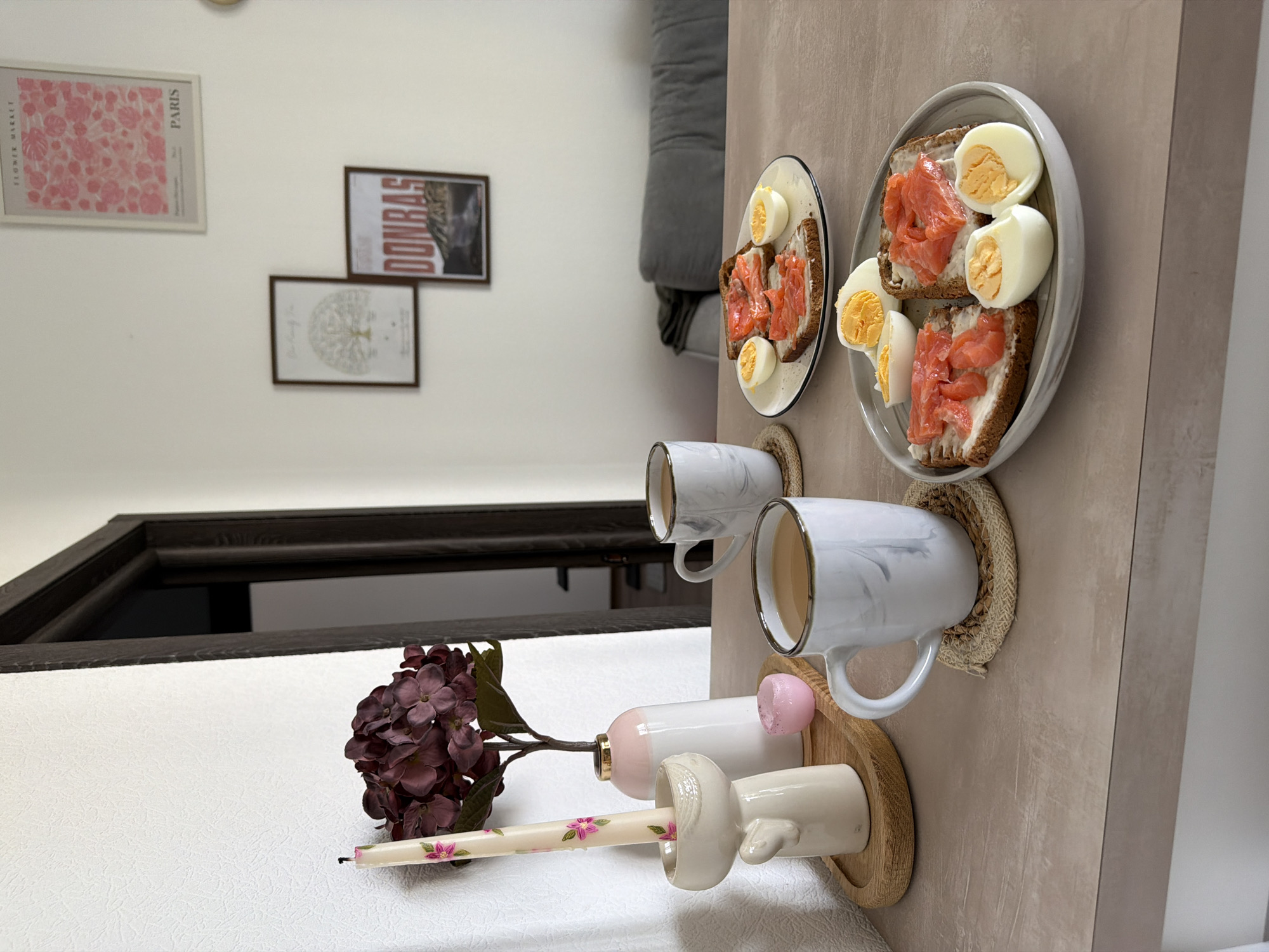 This is a still life photograph taken indoors, likely in an apartment in Kyiv, Ukraine, depicting a breakfast spread on a table. In the foreground, two mugs with a marbled pattern are placed on woven coasters, filled with a light brown liquid, presumably coffee or tea. To the left, a tall, cream-colored candle with floral designs is held upright in a ceramic holder shaped like a cherub. Behind the candle, a vase contains a bouquet of dark purple hydrangea flowers. A small pink decorative object sits near the candle holder.
The table holds two plates with open-faced sandwiches topped with cream cheese and slices of smoked salmon. Each plate also features hard-boiled eggs, cut in half to reveal the yolks. Behind the breakfast spread, a doorway with dark trim leads into another room, partially visible. On the wall in the background, three framed pictures are hung: one with pink floral motifs and the text "PARIS," another with a tree illustration, and a third with red lettering that reads "DONBAS." A blurred grey sofa is visible in the far background. The lighting suggests an indoor setting, possibly during daylight hours, with a diffused natural light source.