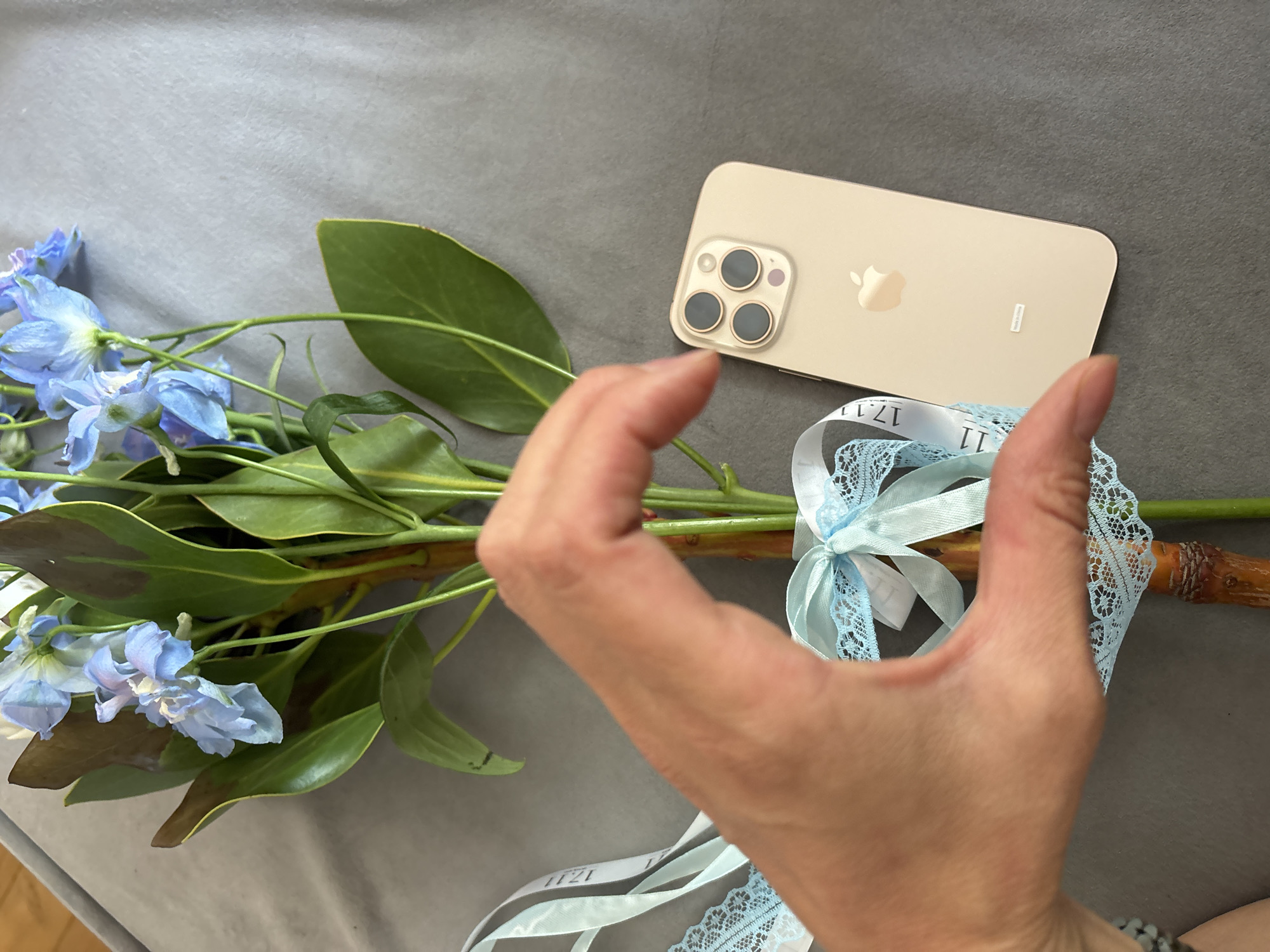 A close-up shot shows a human hand forming a heart shape in front of a bouquet of blue delphinium flowers and green leaves. The bouquet is tied with light blue lace ribbon and a white ribbon with the text "17.11" printed on it. To the right of the flowers and ribbon, a gold iPhone 13 Pro Max is placed on a gray surface. The phone is positioned vertically with the camera array facing upwards. The scene appears to be indoors, on a gray upholstered surface, possibly a couch or bed, and is not located in Chaikovsky, Russia.