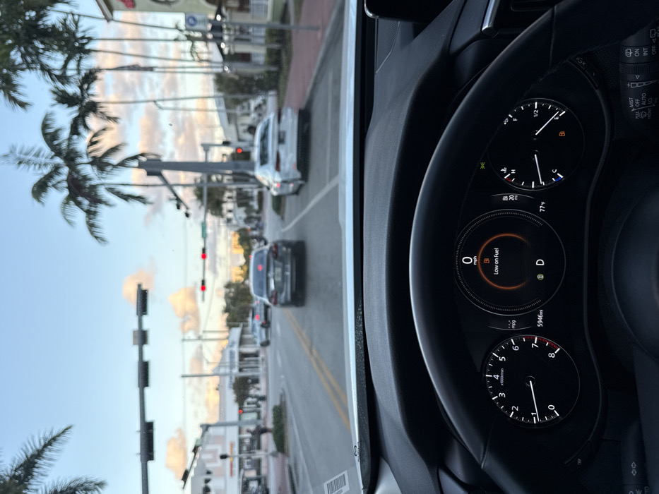 A view from inside a car on a street lined with palm trees, displaying the car's dashboard. The fuel gauge indicates low fuel. The odometer reads 5946 miles. The temperature is 77 degrees Fahrenheit. The car is in drive, with a speed of 0 mph and an engine speed of 3000 rpm. Traffic signals are visible on the street. Other cars are present on the road.