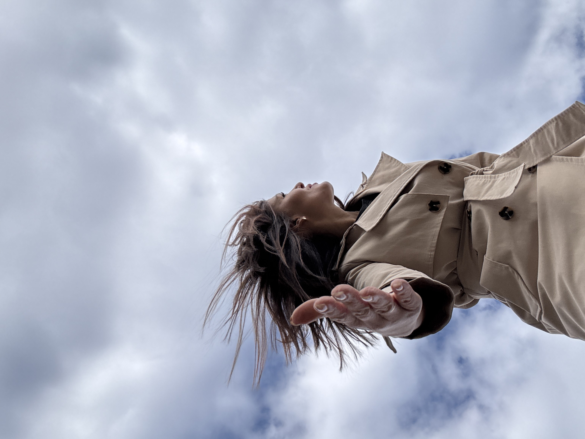 A low-angle shot shows a person in a tan trench coat with their head tilted back looking up at the sky. The sky is filled with white and grey clouds. The person's hand is outstretched towards the viewer. The location is not discernible from the image. There is no indication of the city of Dubovaya, Russia in the photograph.