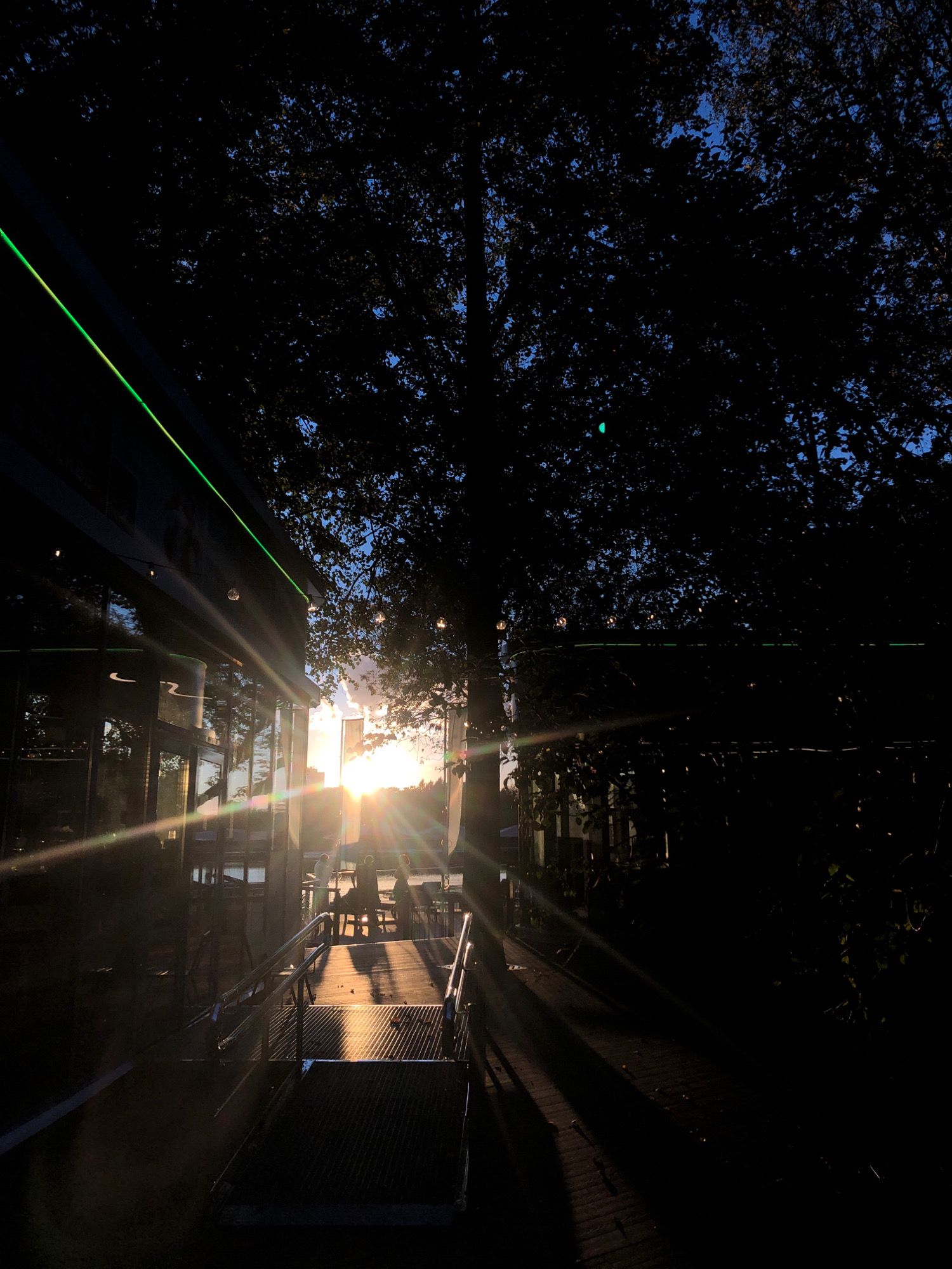 The image depicts a scene at dusk or dawn, with the sun low on the horizon, casting long shadows and lens flares across the frame. The location is identified as Khimki, Russia, likely an outdoor seating area near a building with reflective windows.
In the foreground, there is a metal ramp with handrails leading up to an elevated platform. The ramp has a textured surface. To the left of the ramp, the side of a building is visible, featuring dark reflective windows with a green illuminated stripe running horizontally along its upper edge.
Beyond the ramp, several tables and chairs are set up on a wooden deck, with a body of water visible in the distance. A few silhouetted figures are seated at one of the tables, appearing to be engaged in conversation or enjoying the view.
The background is dominated by the silhouette of a large tree with dense foliage, its branches reaching towards the sky. The sun, partially obscured by the tree and clouds, is a bright, starburst-like source of light, creating prominent sunbeams that radiate outwards and are amplified by lens flares. The sky transitions from a bright orange and yellow near the sun to a deep blue at the top of the frame.
The overall atmosphere is one of a tranquil evening or morning setting, with the natural light creating dramatic visual effects. The presence of the building and outdoor seating suggests a commercial or recreational establishment, possibly a cafe or restaurant.