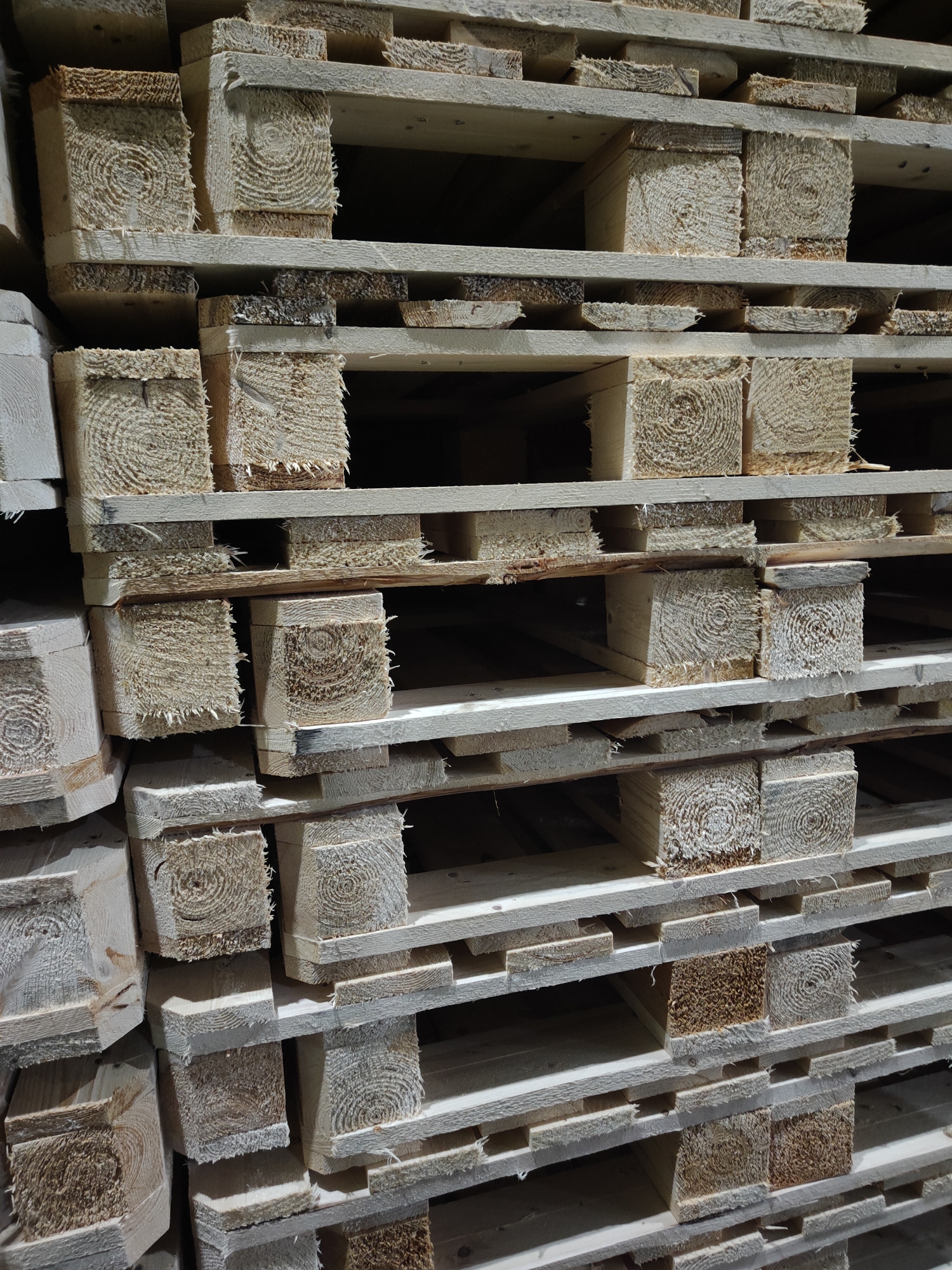 A stack of wooden pallets is shown in a close-up, detailed shot. The pallets are made of light-colored wood and are stacked in multiple rows. Each pallet consists of horizontal planks supported by vertical blocks. The wood grain is visible, with circular patterns on the ends of the blocks. Some of the wood appears rough and unfinished, with splintered edges on the blocks. The lighting creates shadows between the pallets, highlighting the depth and texture of the stacked items. The background is dark and indistinct, suggesting an indoor or shaded storage area. There are no people or animals visible in the image.