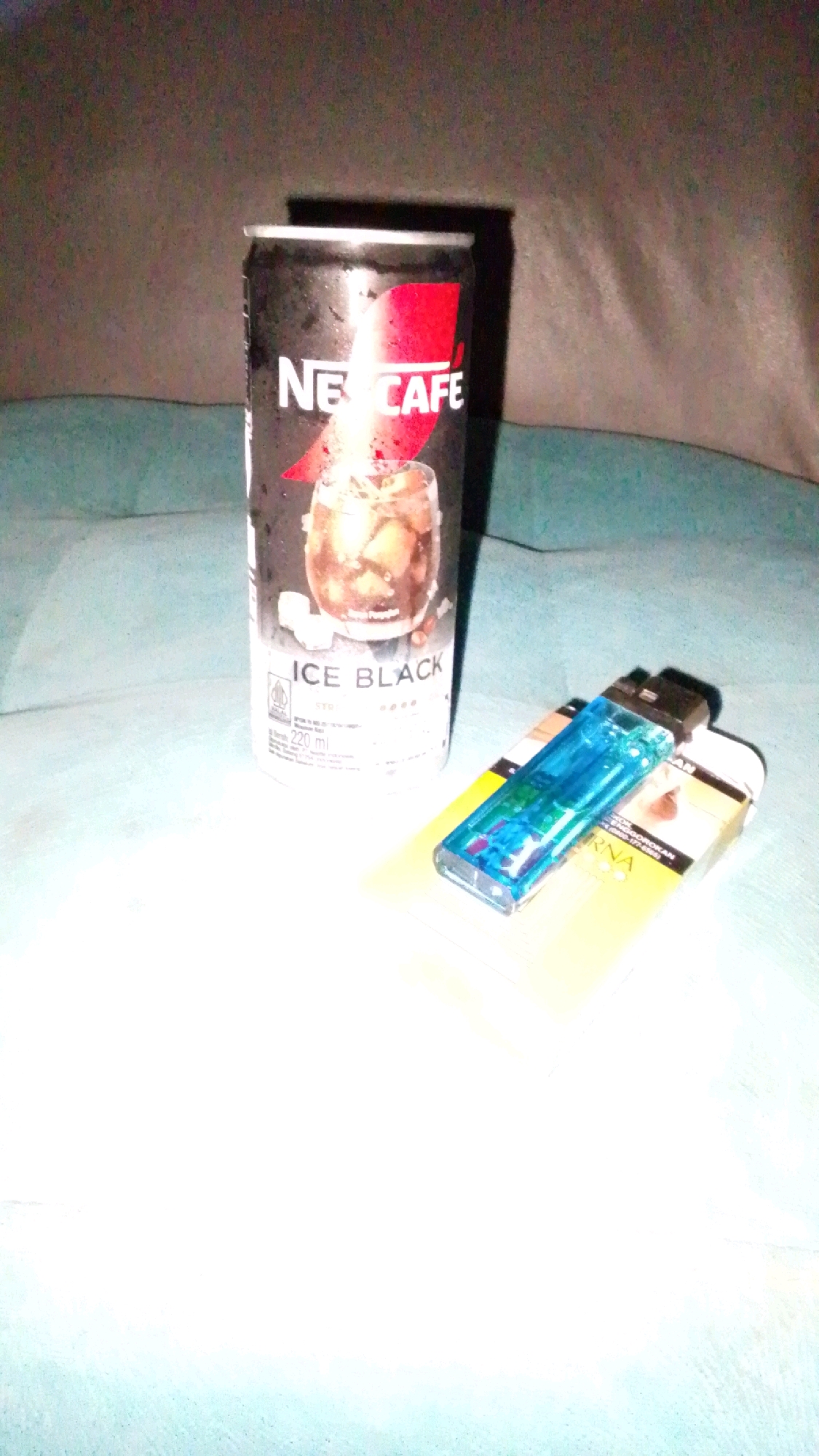 A Nescafé Ice Black can and a blue lighter are placed on top of a pack of cigarettes. The Nescafé can is black with a red logo and an image of a glass of iced coffee. The pack of cigarettes is yellow with some text in Indonesian. The scene appears to be indoors, possibly on a bed or couch, with a dark, textured background. The lighting is dim, and there is a strong glare on the Nescafé can. There is no indication of the location being specifically in Cintaratu, Indonesia, beyond the potential origin of the products.