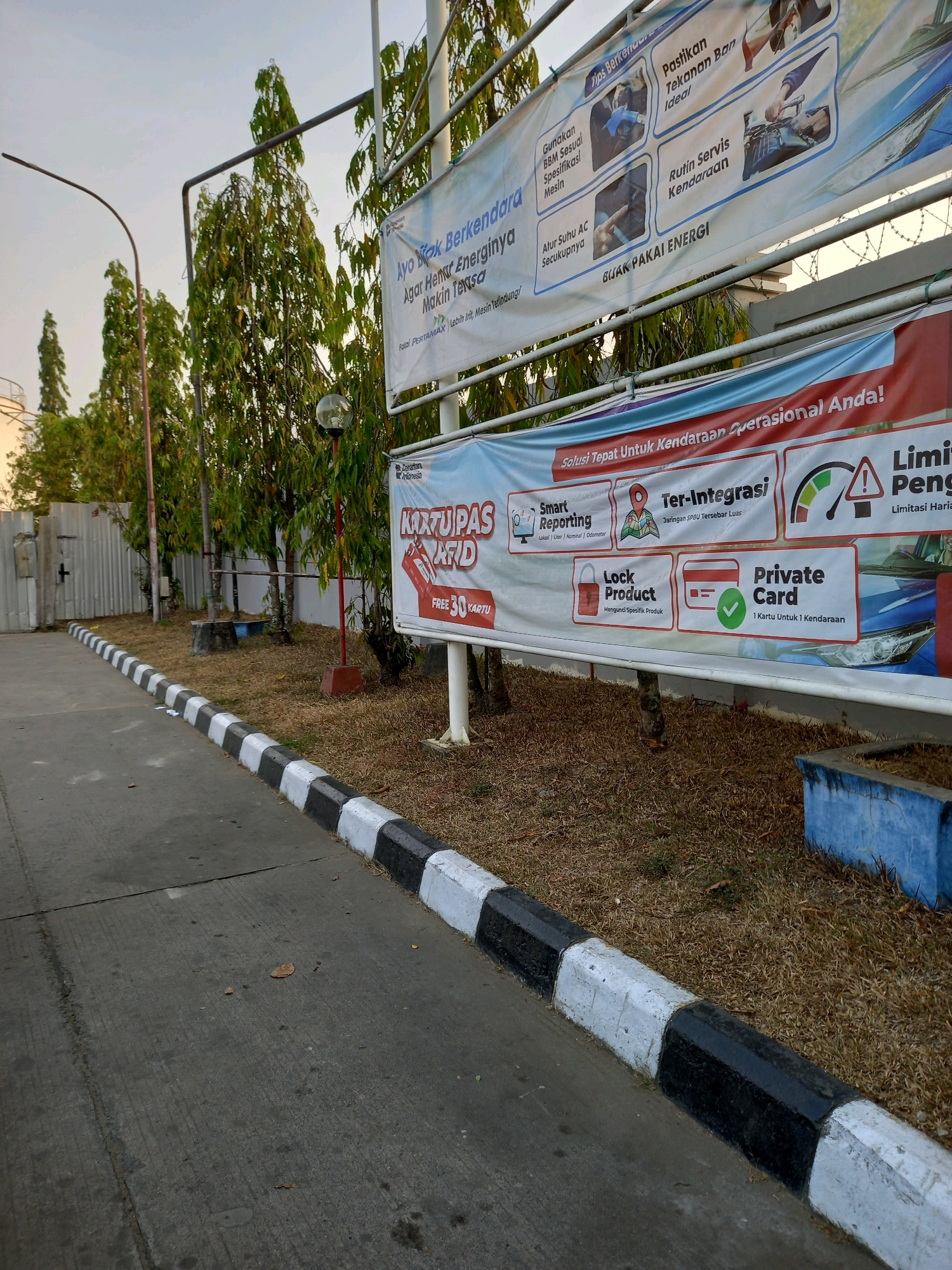 The image depicts a street scene in Samarinda, Indonesia, likely near a fuel station. In the foreground, a paved road with a black and white striped curb runs along the right side. Beyond the curb, dry grass and some vegetation are visible. Several trees with green foliage stand tall behind a metal fence. A street lamp is present to the left. In the middle ground, two large banners are displayed horizontally. The top banner, mostly white, has text in Indonesian and illustrations related to vehicle maintenance and fuel efficiency, including tips on fuel usage, tire pressure, and engine specifications. The bottom banner, predominantly red and white, features text in Indonesian promoting a "Smart RFID Card" for vehicles, with icons representing reporting, integration, and card features. The overall environment suggests an outdoor setting, possibly during the late afternoon or early evening, given the lighting. There are no people visible in the frame.