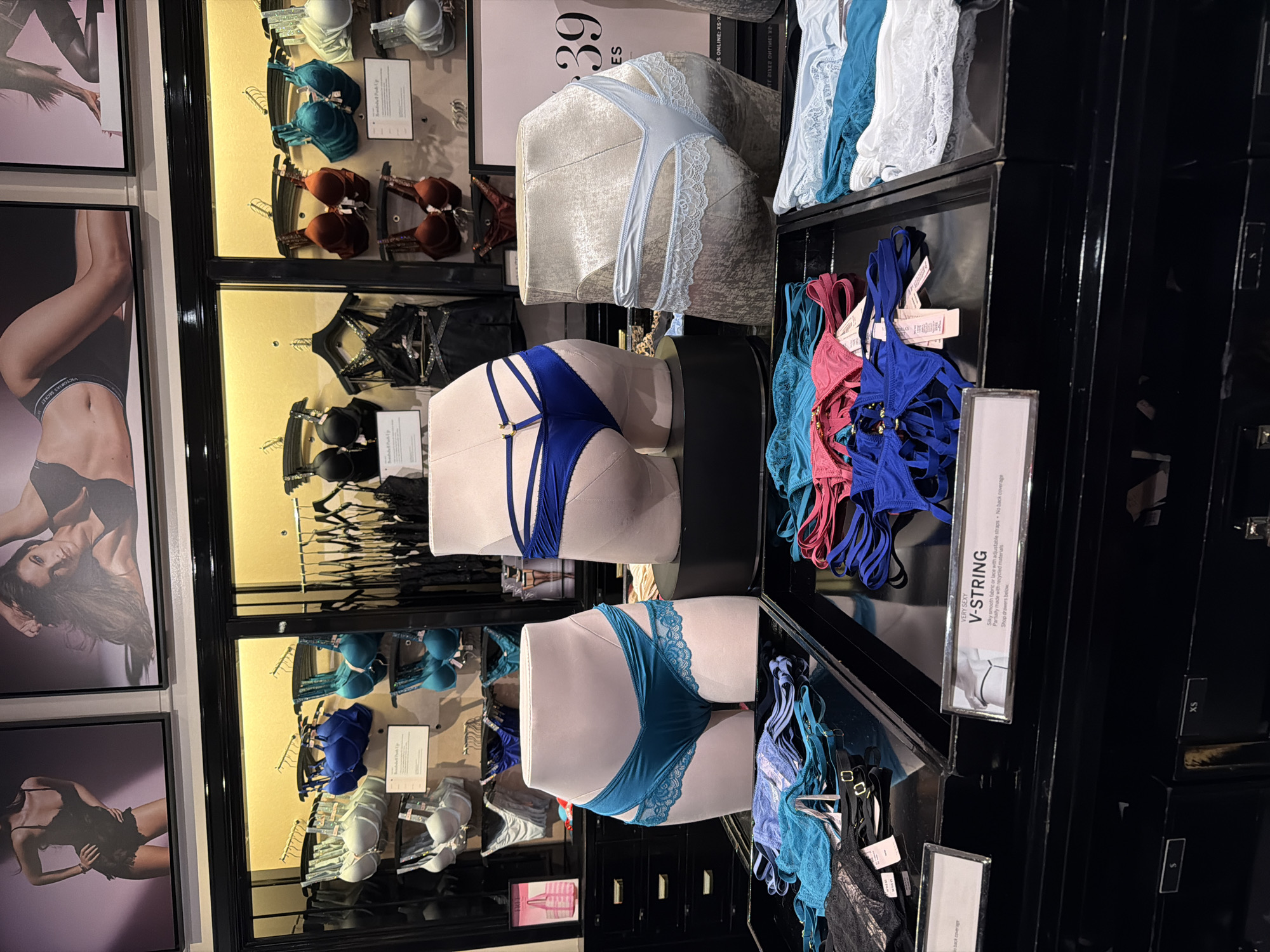 This media file captures a retail display of lingerie in a store, likely in New York, United States of America. The scene is brightly lit, suggesting it's during business hours. The display features various types of lingerie, including bras, panties, and thongs, on mannequins and hangers. The items are predominantly in shades of blue, teal, pink, and white, with some black lace accents.
Notable details include a mannequin showcasing a light blue lace thong and another displaying a blue strappy brief. Several framed images of models wearing lingerie are visible on the left side of the display. A sign on the right side reads "VERY SEXY V-STRING" and provides details about the product's material and features. Other visible text includes sizing information like "XS" and "S," and a partial website address. The overall impression is a well-organized and visually appealing lingerie department within a retail environment. There are no clear indications of the time of day or weather. The focus is on the merchandise and its presentation.