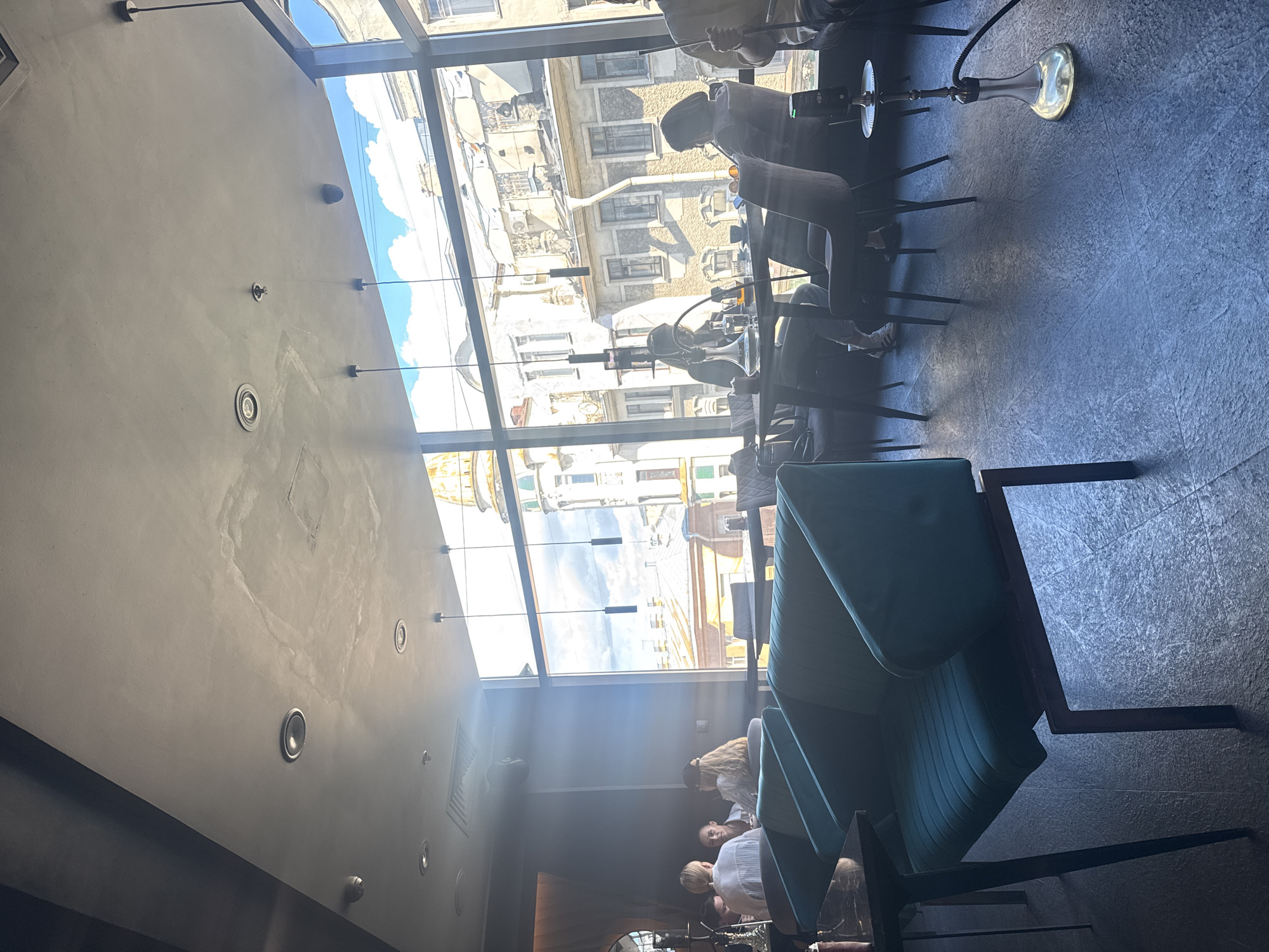 The image depicts the interior of a modern hookah lounge with large windows offering a view of an urban street scene. Several people are seated at tables, some engaged in conversations and others using hookahs. The furniture includes plush teal velvet sofas and chairs, with dark marble-topped tables. The lighting is a mix of natural light streaming through the windows and artificial lighting from pendant lamps. Outside, the architecture suggests an older European city with ornate buildings and balconies. A hookah apparatus with a silver base and black hose is visible on the floor in the foreground.