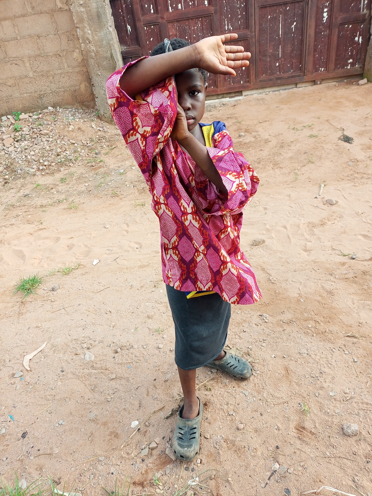 A young person stands on sandy ground, shielding their face from the camera with their left hand. They are wearing a colorful, patterned shirt with pink and red designs and a dark skirt. Dark, worn clogs are on their feet. Behind them is a rustic, wooden gate and a brick wall. The environment appears to be an outdoor, unpaved area, consistent with a location in Tempure, Nigeria.
