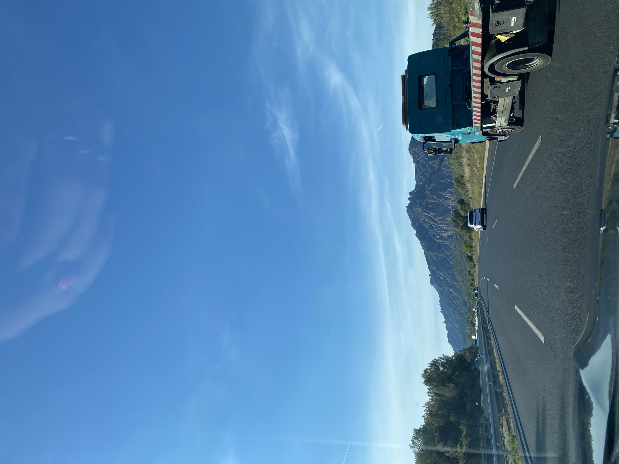 A slightly tilted, broad shot captures a clear blue sky over a highway, reflecting in the car window on the left. The highway stretches into the distance, lined with guardrails and scattered vegetation on one side. A tow truck is positioned on the right of the frame, followed by a mountain range in the background. The location appears to be along a road near Esparreguera, Spain, suggested by the mountainous terrain in the distance.