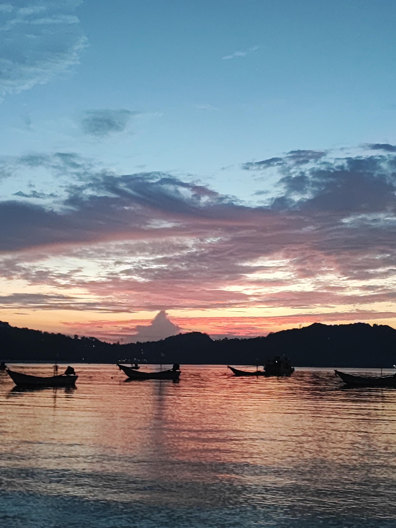 This is a photograph of a serene sunset scene over the water near Ko Pha-ngan District, Thailand. The sky is a gradient of soft blues at the top, transitioning into a vibrant mix of pinks, oranges, and yellows as it nears the horizon. Wispy clouds are scattered across the sky, catching the colorful hues of the setting sun.
In the foreground, the calm water reflects the colors of the sky, creating a beautiful mirror effect. Several small, dark silhouettes of boats are scattered across the water. These appear to be traditional fishing boats, some with a single mast or motor. They are at rest, bobbing gently on the waves.
The background features the dark, silhouetted outline of a hilly coastline. The details of the land are obscured by the low light of dusk, but the overall shape suggests lush vegetation. A single, distinct cloud formation rises from the silhouette of the land, resembling a small, pointed mountain.
The time of day is clearly sunset, indicated by the rich, warm colors in the sky and their reflection on the water. The weather appears calm and peaceful, with smooth water and gentle clouds. The scene evokes a sense of tranquility and natural beauty. There is no visible text in the image.