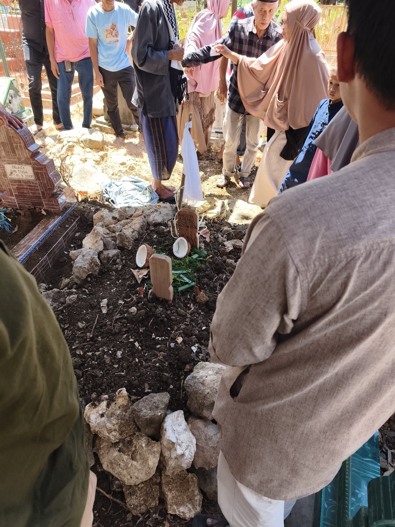 This image captures a solemn scene at a cemetery in Tonasa, Indonesia, likely a burial site. Several individuals are gathered around a grave, some in traditional attire. The grave itself is a disturbed mound of earth, adorned with stones and a wooden marker bearing an inscription, possibly a name and dates. Nearby, two halves of a coconut lie on the ground, a common offering at Indonesian graves. The presence of multiple people and the grave decorations suggest a recent burial or memorial service, evoking a somber atmosphere within the outdoor environment.