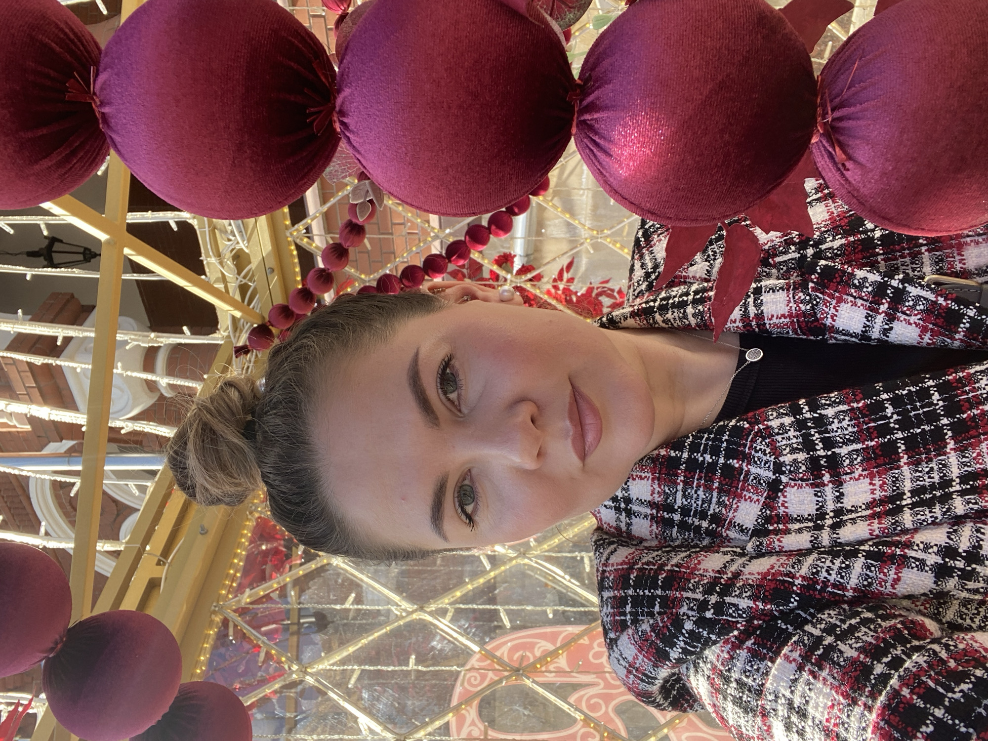 The image shows a woman in Izhevsk, Russia, positioned in front of a Christmas display. The display consists of red decorative balls and decorative lighting. The woman has her hair in a bun and is wearing a black top with a red, black, and white plaid jacket.