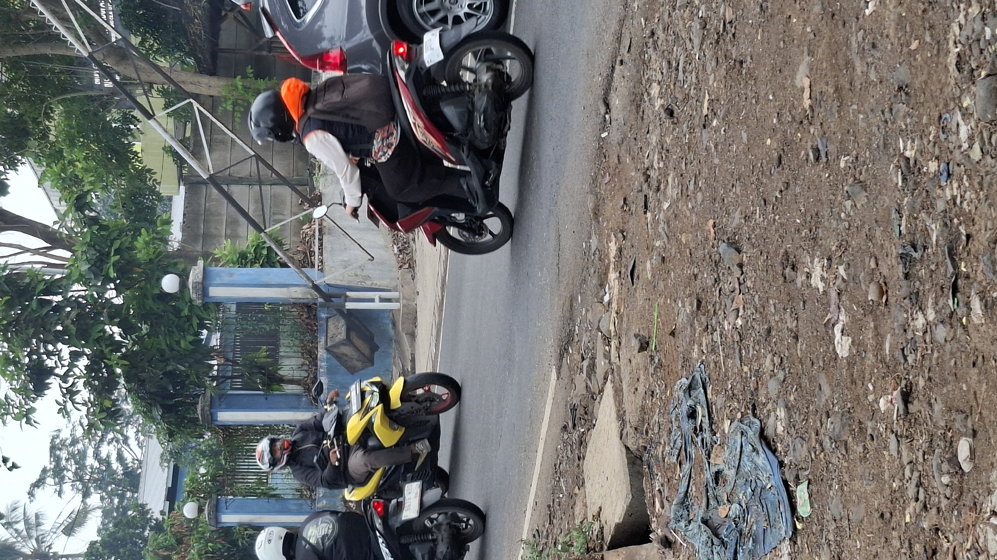 In Cilaku, Indonesia, on a cloudy day, two motorcycles and one car are traveling on a paved road. The motorcycle on the left is black, and the rider is wearing a black jacket and helmet. The yellow motorcycle in the middle is ridden by a person wearing a black jacket and a helmet with an orange chin strap. The red motorcycle on the right is being ridden by a person in a white long-sleeved shirt, a dark vest, and an orange hood. This rider is also wearing a black helmet and has a brown backpack. A gray car is visible behind the red motorcycle. The roadside features a blue and white gate with decorative posts topped by white spheres, and beyond it, a fence and trees. The ground on the right side of the road is a mix of dirt and rocks, with some scattered debris, including a large piece of blue tarp.