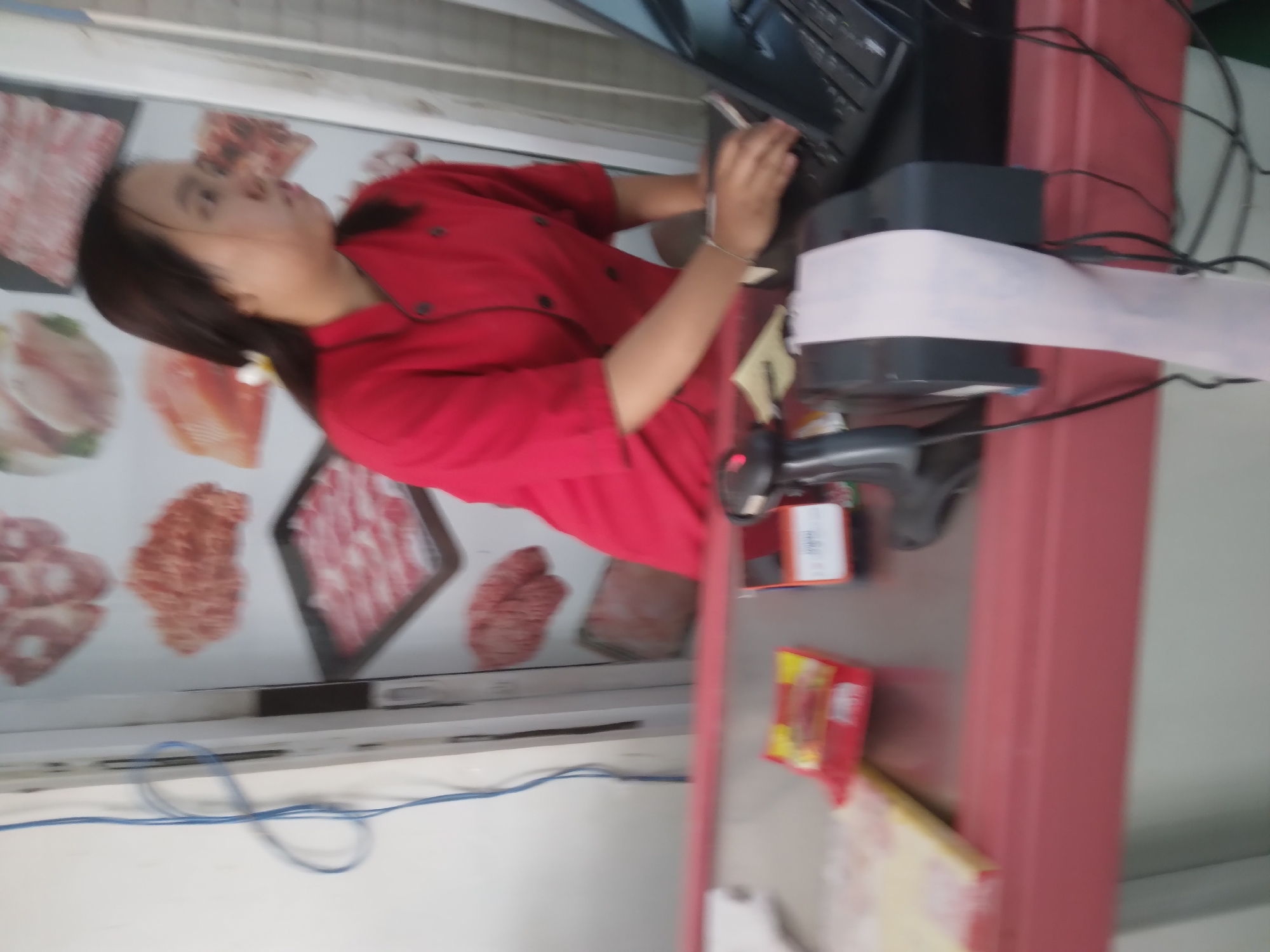 A young woman wearing a red chef's jacket with black trim is standing behind a red counter, using a laptop. She has dark hair and is looking to the right. On the counter, there is a receipt printer with a long pink receipt, a scanner, and some packaged goods. Behind her, there are posters of meat products on a white wall. The overall scene appears to be a food establishment, possibly a butcher shop or a restaurant, located in Sokaraja, Indonesia.
