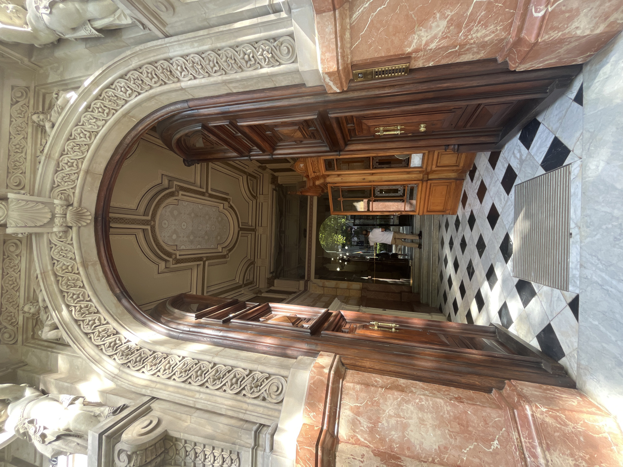 This image shows the ornate entrance to a building in Barcelona, Spain. The scene is viewed from a low angle, looking through an open, grand doorway. The doorway itself is constructed of dark, polished wood with intricate carvings and brass handles. Above the doorway, a large, arched stone structure features detailed sculptural elements and decorative moldings. The archway leads into a dimly lit hallway with a checkered black and white marble floor. Sunlight streams in from the right, casting shadows and highlighting the textures of the marble and stone.
To the right of the doorway, a wall is clad in striking red and white veined marble. Above this marble, a brass intercom panel with numerous buttons is visible. Further down the hallway, a wooden cabinet with glass doors stands against the wall. In the distance, a person wearing a white shirt and beige pants is visible, walking away from the camera. They appear to be entering or exiting the building. The overall impression is one of grandeur and historical architecture. The lighting suggests it is daytime, possibly late morning or early afternoon given the angle of the sunlight.