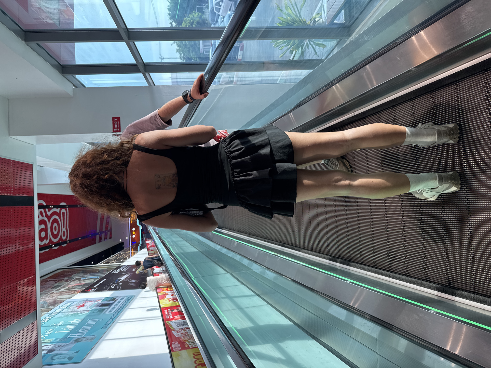 An adult woman with curly brown hair is standing on an escalator in a public building, possibly a mall, in Đông Hoa Lư Ward, Vietnam. She is facing away from the camera, wearing a black tank top, a black ruffled skirt, white socks, and grey sneakers. A man, partially visible, is standing next to her on the escalator, holding the handrail. The escalator is equipped with green lighting along its side. In the background, there are advertisements and windows revealing an outdoor scene with vehicles. The overall environment appears bright and modern, with a focus on commercial activity.