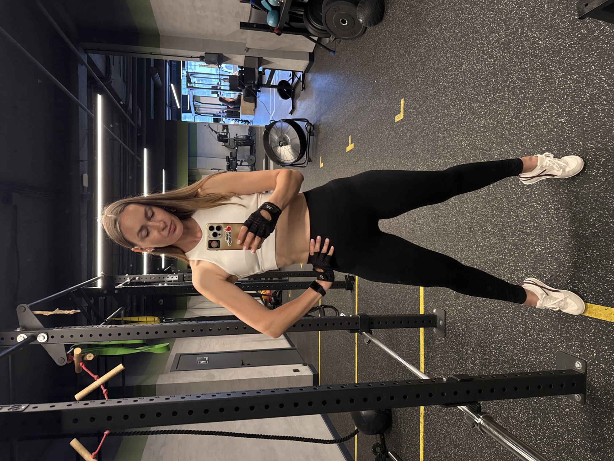 A woman with long blonde hair is posing for a selfie in a gym. She is wearing a white tank top, black leggings, and white sneakers. She is also wearing black fingerless gloves and a fitness tracker on her left wrist. The gym has a rubberized floor with yellow lines. There are weightlifting racks and other gym equipment in the background. A large fan is visible in the middle ground. The lighting in the gym is bright and modern, with overhead lights and strip lighting on the walls. The scene appears to be taking place indoors during the day. The woman has a slight smile on her face, suggesting she is feeling confident and happy. There is text visible on her phone case that says "BRUNO CIRO" and some Cyrillic text.