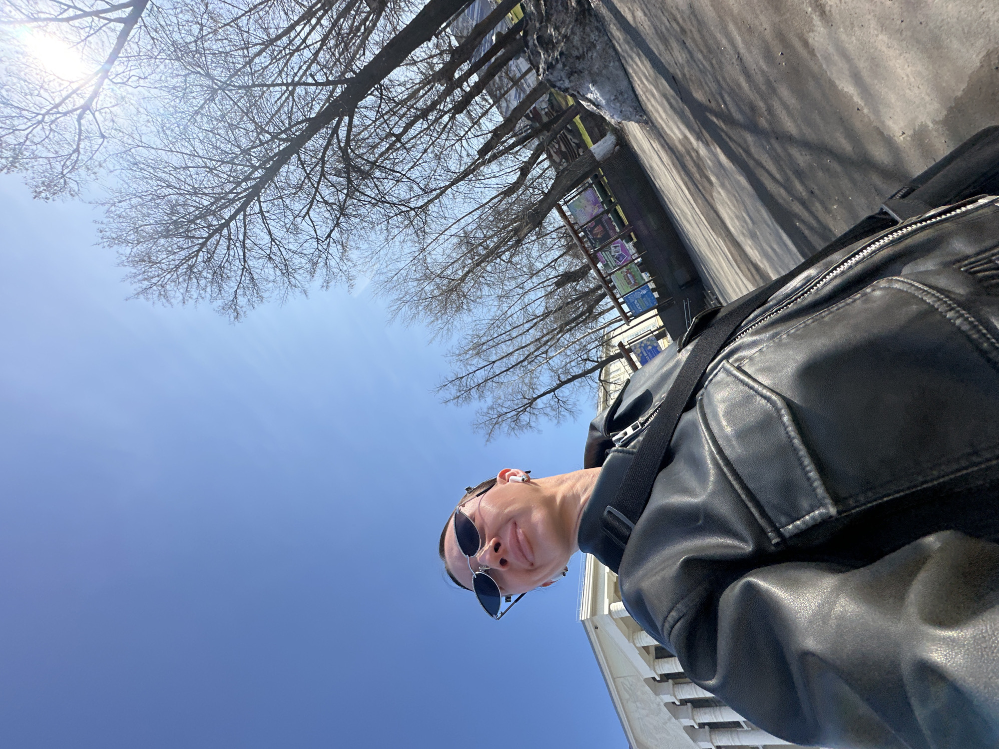 A tilted outdoor selfie features a young person in the lower right, looking up towards the camera with a slight smile. The person wears black sunglasses, a white earpiece in their right ear, and a black leather jacket with a black strap from a bag visible across their shoulder.
The upper and central portions of the image are dominated by a clear, bright blue sky, against which numerous bare tree branches and trunks are silhouetted. A strong sun flare emanates from the upper left, indicating a sunny day, likely mid-morning or afternoon, as evidenced by the long shadows cast across the paved ground visible on the right side of the frame. In the distance, beneath the trees and alongside the paved area, structures featuring colorful panels, possibly advertisements or public art, can be seen. A light-colored building facade is partially visible in the lower right background. The scene suggests an urban or park setting in Votkinsk, Russia, during a season without foliage, likely late winter or early spring. No discernible text is visible.