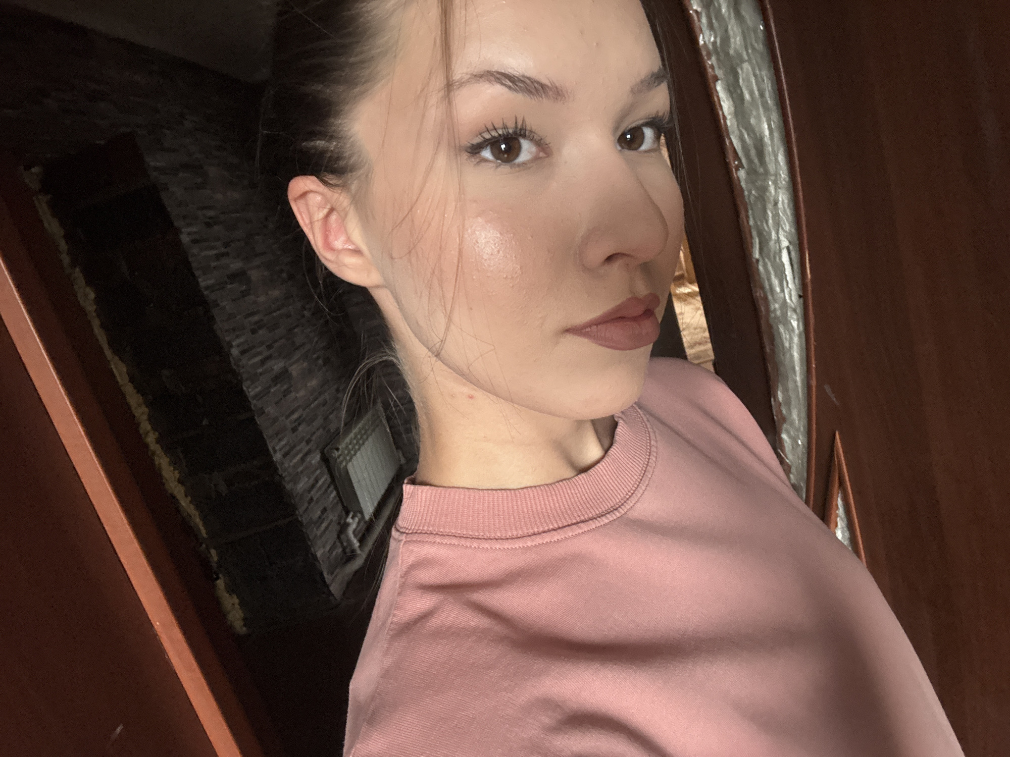 The image is a close-up, high-angle selfie of a young woman with brown hair pulled back, taken indoors. She is wearing a pink crewneck sweatshirt. Her face is turned slightly to the right, and she is looking towards the camera. Her makeup includes mascara and lip color. Her skin appears dewy. The background is dark and appears to be a hallway with exposed brick on the left and a wooden door or frame on the right. There is a radiator visible on the brick wall. The location in Vishnevka, Russia, is not discernible from the image itself.