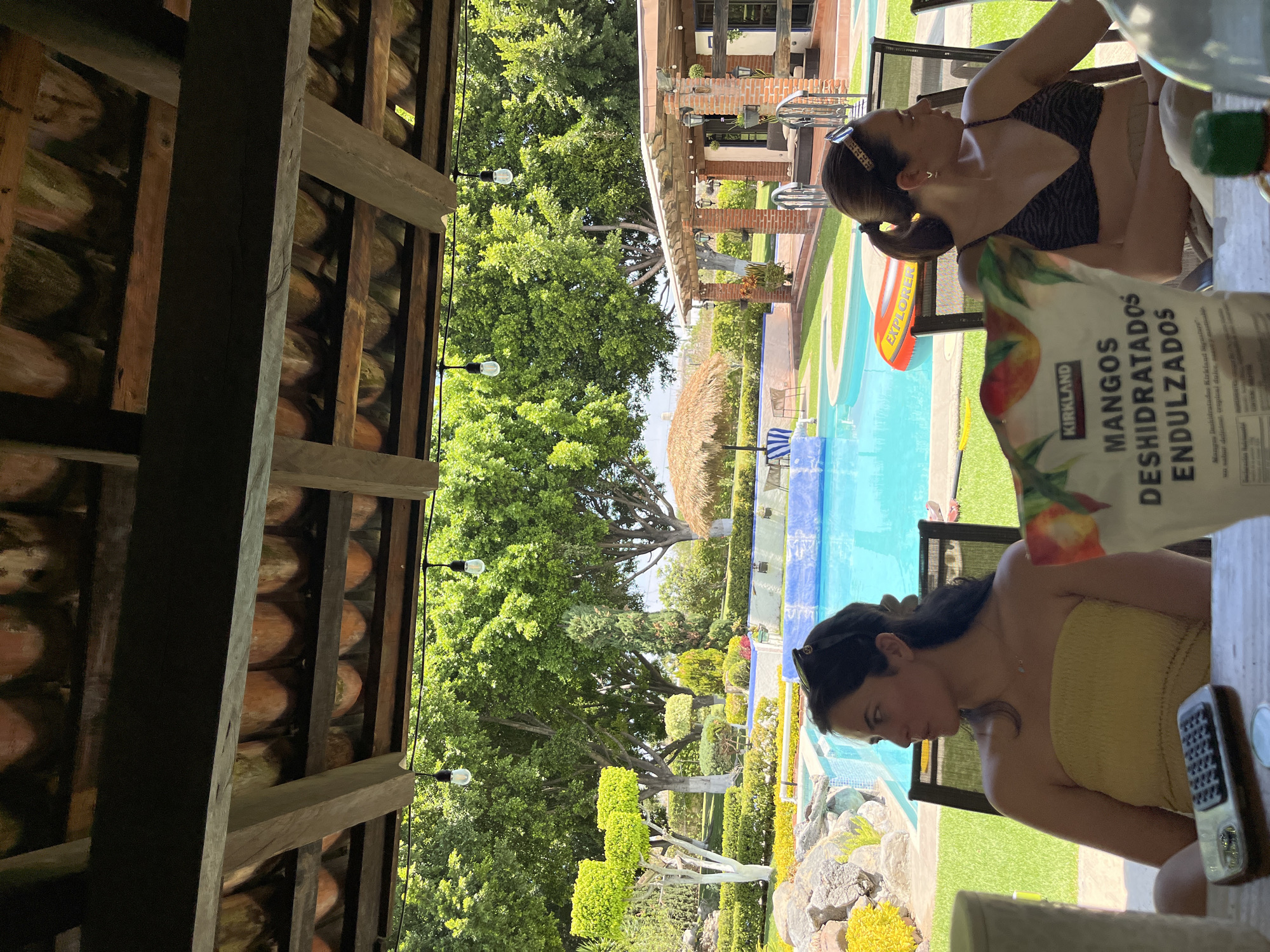 The image shows two women seated at an outdoor table by a pool. A bag of Kirkland dried mangoes is visible on the table. The pool is surrounded by well-manicured landscaping, including trees, shrubs, and flowers. A thatched umbrella is also visible. The women are in Unknown, Unknown.