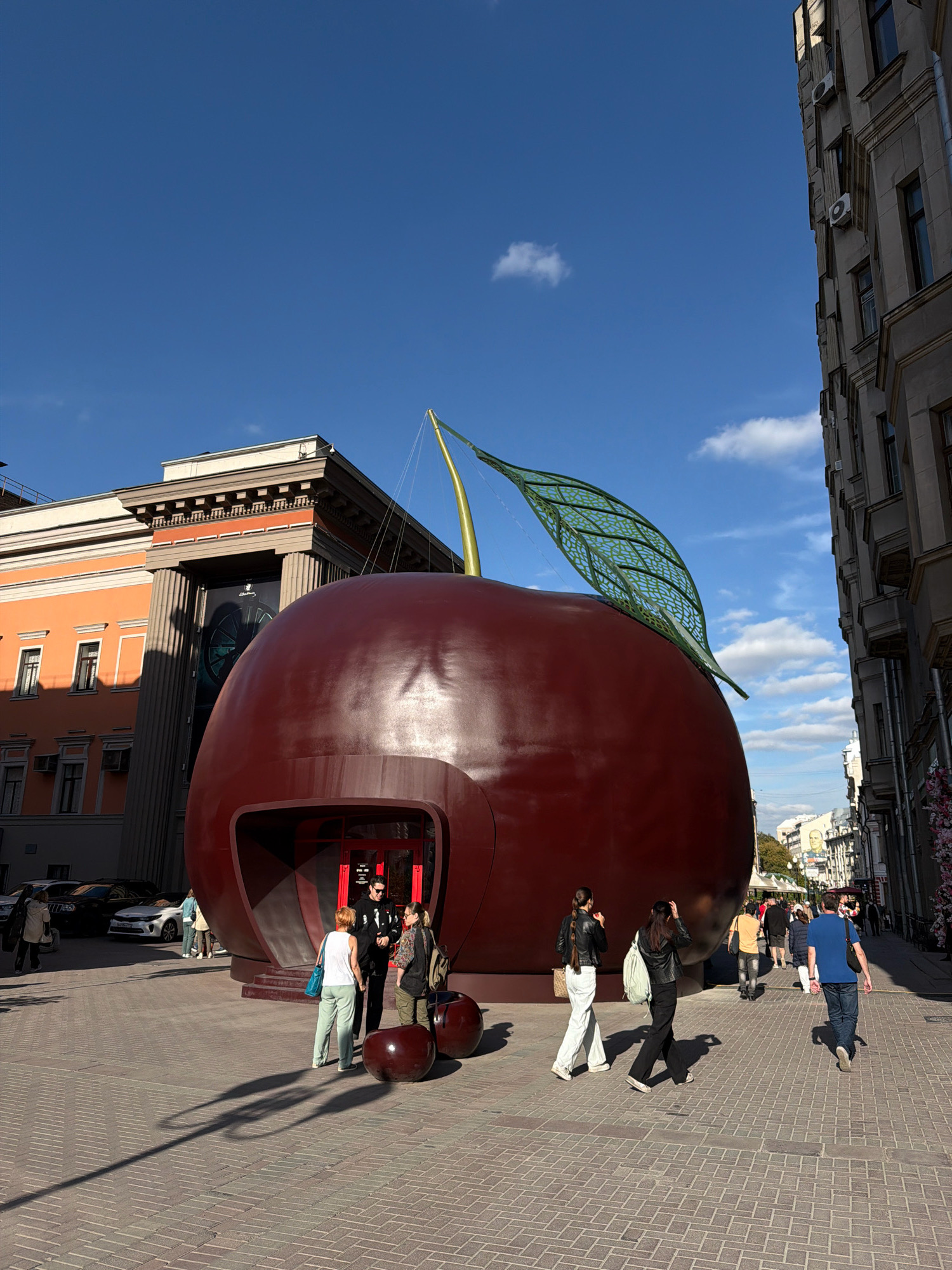 The image depicts a sunny day in Moscow, Russia, with a bright blue sky and scattered clouds. In the foreground, a massive, glossy red cherry sculpture dominates the scene, featuring a stylized green leaf and stem. The sculpture has an opening resembling a doorway, with steps leading up to it.
Several people are visible in and around the sculpture. Three individuals stand near the entrance, two men and one woman. One man wears a black leather jacket and sunglasses. Another woman, in the foreground, has blonde hair and wears a white tank top and green pants. A man in a black jacket and sunglasses is positioned behind her.
Further out in the plaza, which is paved with brickwork, several other individuals are walking in different directions. Some are dressed casually in jackets and pants. The sun casts long shadows from the people and the sculpture onto the ground.
In the background, to the left, an orange building with classical architectural elements stands tall, featuring columns and ornate detailing. To the right, a multi-story building with a grey facade and numerous windows lines the street. Cars are parked along the street in the distance. A large banner with a circular graphic is visible on the facade of the orange building. The overall atmosphere suggests a bustling urban environment on a pleasant day.