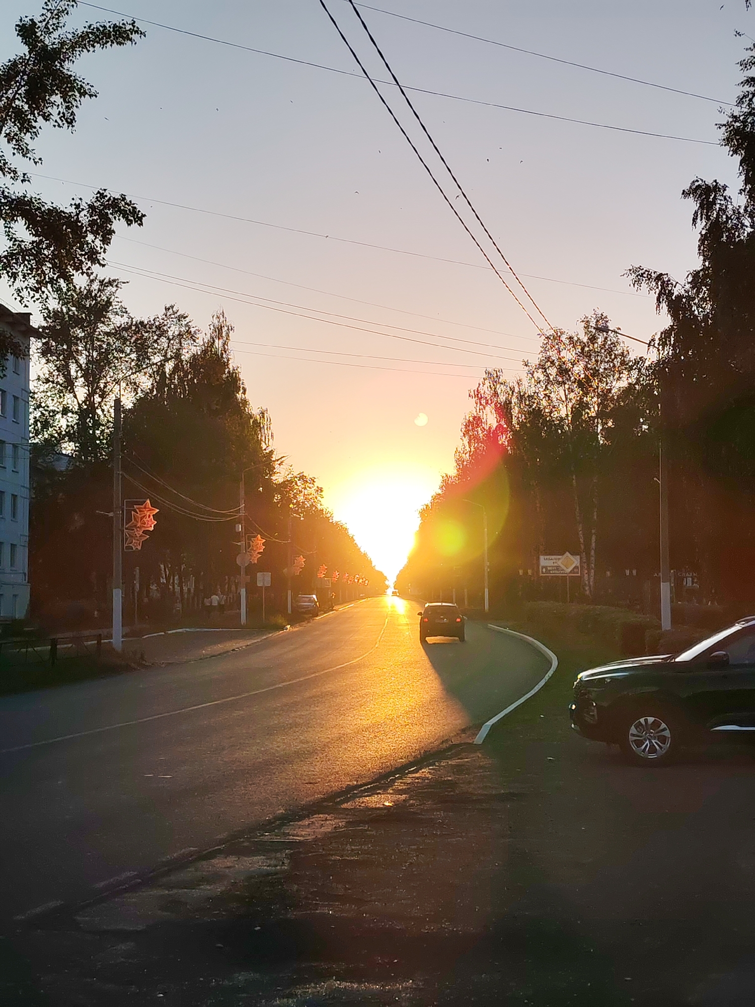 The image depicts a street scene in Chaikovsky, Russia, during a sunset. The sun is low on the horizon, casting a warm, golden light across the asphalt road and the surrounding trees. The sky is a gradient of pale blue and soft pink.
On the left side of the road, a multi-story residential building is visible, with several windows. Adjacent to the building and lining the street are numerous trees, mostly deciduous, with some birch trees interspersed. Decorative star-shaped lights are mounted on poles along the sidewalk, some illuminated by the setting sun.
Two cars are present on the road. One dark-colored car is in the foreground on the right, parked on the shoulder. Another dark-colored car is further down the road, driving away from the viewer. A third car is visible further up the road, also driving away.
A signpost with Cyrillic lettering is visible on the right side of the road, amidst the trees. Overhead, power lines stretch across the sky. The road surface shows some wear and tear, with patches and a distinct lane marking. The overall impression is a peaceful, urban landscape illuminated by the dramatic light of sunset.