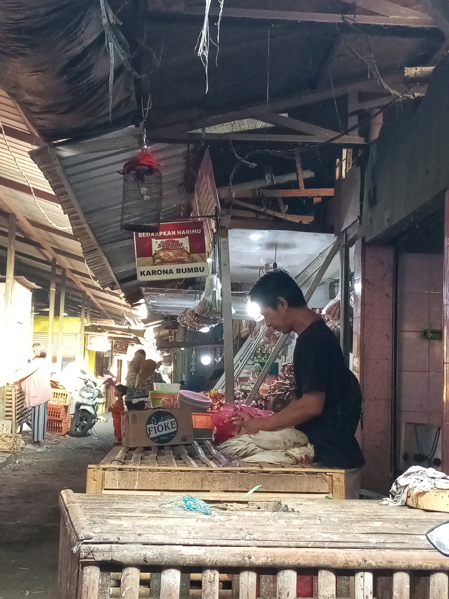 In Setiamekar, Indonesia, a man in a black t-shirt is standing at a wooden stall in an outdoor market. He is in the process of preparing a white chicken, which is lying on a flat surface in front of him. The market appears to be covered by a corrugated metal roof, with some areas draped with dark fabric. Various goods are visible in the background, including crates, a motorcycle, and other market stalls with awnings and hanging items. There are other people in the background, including a child and a woman wearing a headscarf, as well as some customers examining products. A sign advertising "KARONA BUMBU" and "Mie Sedaap" is hanging from the roof.