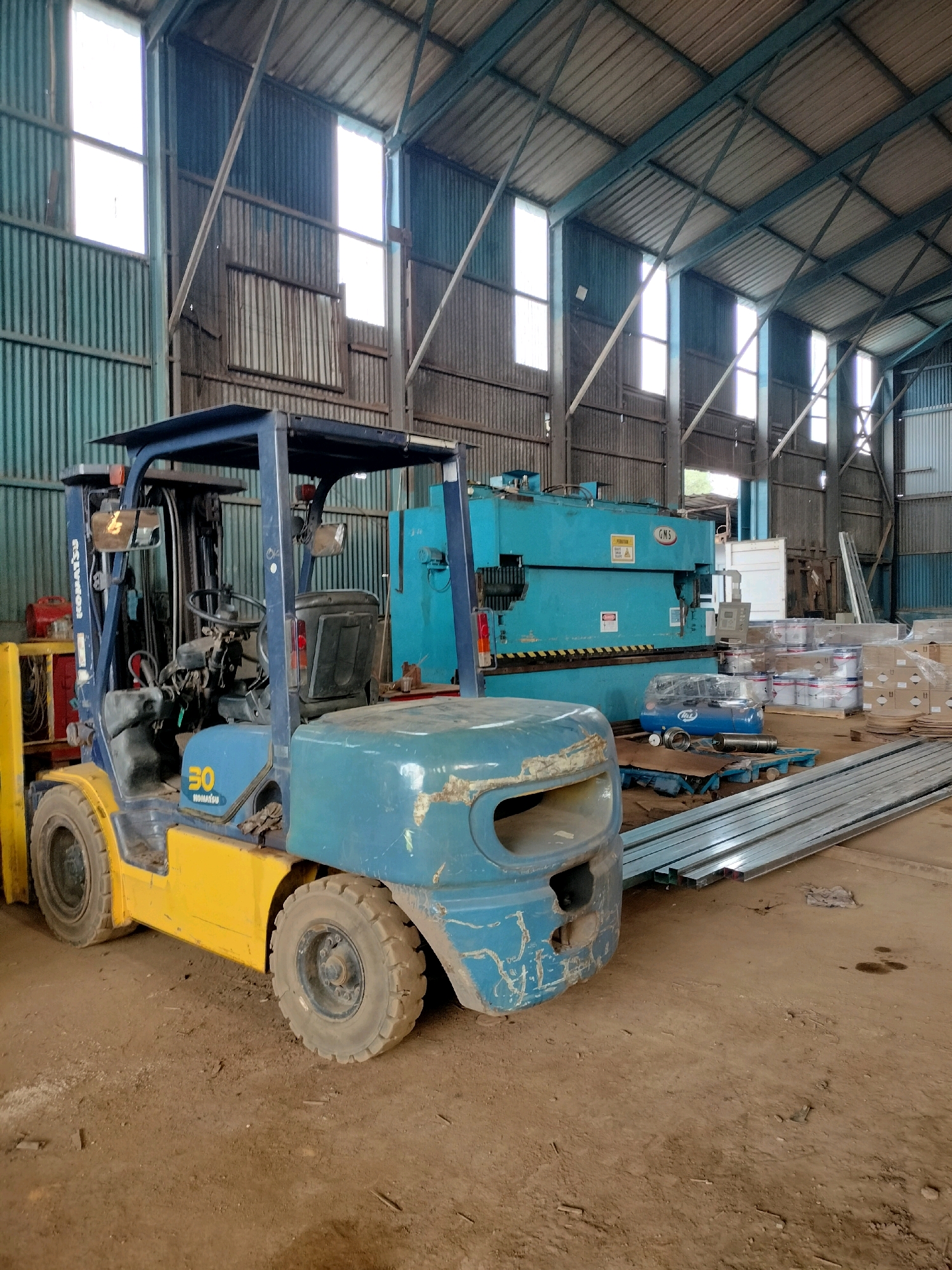 This image depicts the interior of an industrial warehouse located in Batam, Indonesia. The structure features a corrugated metal roof supported by steel beams and corrugated metal walls. Natural light filters in through rectangular windows spaced along the upper sections of the walls.
The foreground is dominated by a blue and yellow Komatsu forklift. The forklift is parked on a dirt floor and has visible wear and tear. Adjacent to the forklift, to its right, is a stack of long, thin metal beams.
In the mid-ground, a large, teal-colored industrial machine, possibly a press brake, is situated. To the right of this machine, several stacks of cylindrical containers, likely paint cans, are arranged on pallets. A smaller, blue air compressor is visible near the industrial machine. A red fuel can is also present near the forklift.
The environment appears to be an active industrial workspace, with tools and equipment characteristic of a manufacturing or fabrication facility. No people are visible in the image.