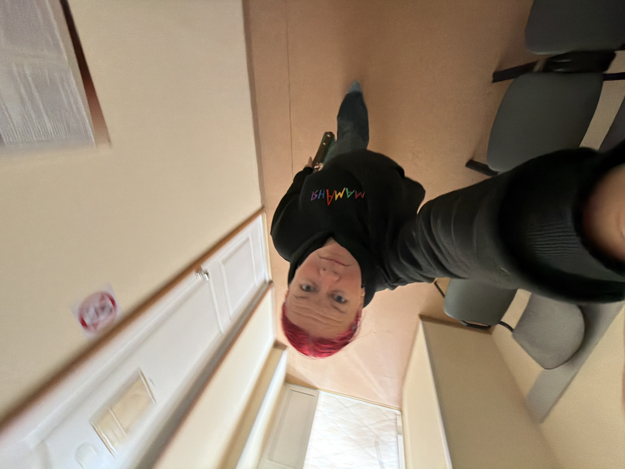 The image, captured from a high-angle perspective, shows a person indoors taking a selfie. The individual, positioned centrally and looking directly at the camera, has short, vibrant pink/red hair, light skin, and blue eyes, displaying a neutral expression. They are wearing a black hooded sweatshirt adorned with colorful embroidered text and dark trousers or jeans, complemented by light blue socks.
The visible text on the sweatshirt, spelled out in rainbow colors, reads "MAMAHЯ" (M A M A H Я), which appears to be Cyrillic script.
The setting is an indoor environment featuring light beige/cream walls and a light brown/tan floor. On the right side of the frame, several chairs with gray cushions and black frames are arranged. To the left, a white paneled door is visible. Attached to the wall near the door is a small, circular red and white sticker or sign, with a transparent rectangular object positioned below it. A window or reflective surface is partially visible towards the bottom of the frame.
The image is significantly rotated, creating an unusual perspective where the floor appears as a wall. The lighting suggests an indoor setting, likely with artificial illumination, as no specific cues for the time of day or weather are discernible. While the location context of Chaikovsky, Russia, is provided, the image itself does not contain any visual elements to confirm this specific geographical location. The primary activity depicted is the person taking their own photograph.