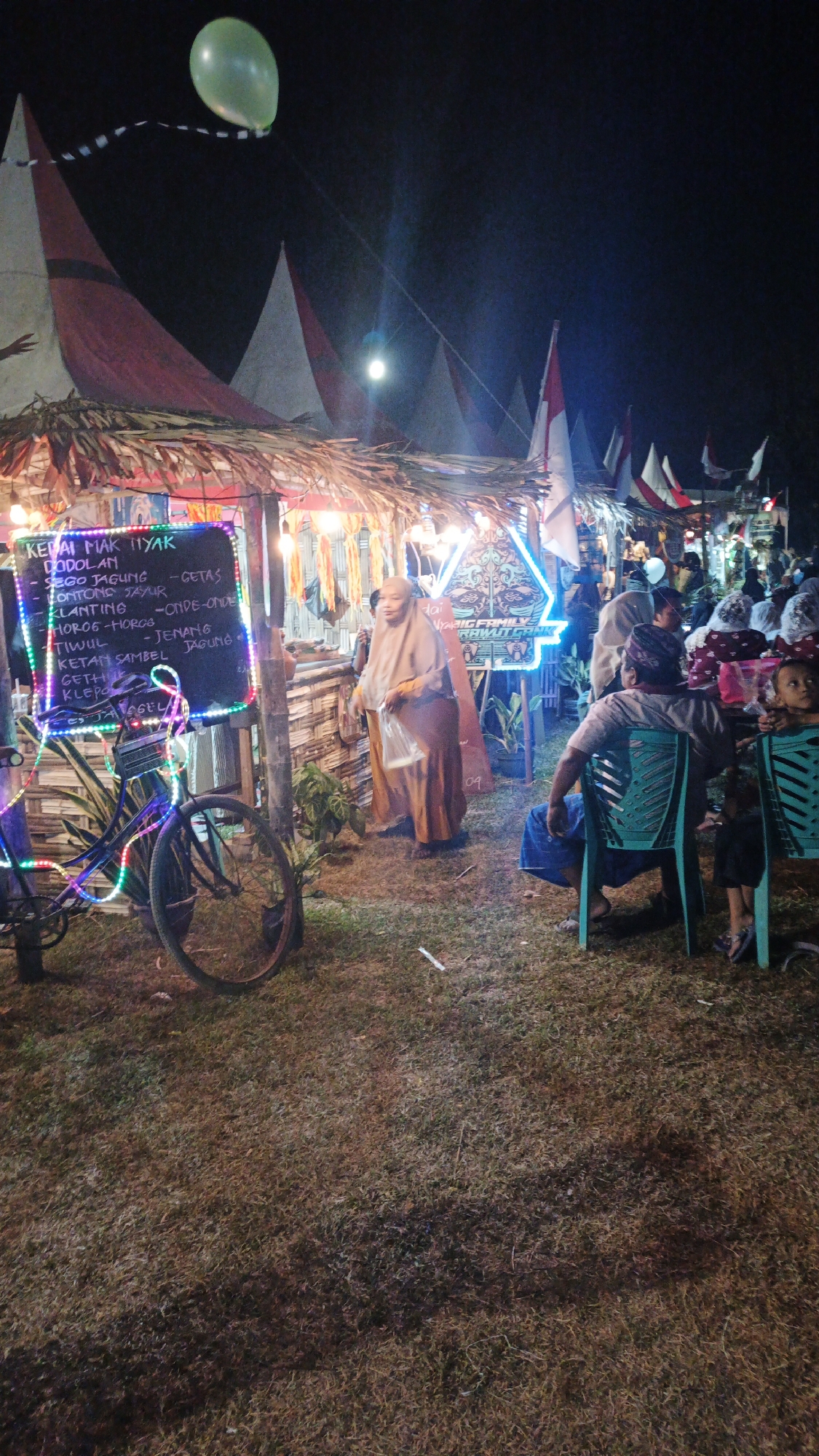 The image depicts an outdoor night market scene in Ketemasdungus, Indonesia. In the foreground, a bicycle decorated with colorful LED lights is parked. To the left of the bicycle, a blackboard menu is displayed, listing various food items like "DODOLAM," "SEGO JAGUNG - GETAS," "LONTONG JAYUR," "KLANTING - ONDE-ONDE," "HOROG-HOROG," "TIWUUL - JENANG JAGUNG," "KETAN SAMBEL," and "GETHI." A woman wearing a tan hijab and a long tan dress is standing near the stall, holding a plastic bag. In the background, several stalls are visible, adorned with Indonesian flags and lights, suggesting a festive atmosphere. People are gathered around the stalls, and some are seated on chairs, indicating a lively market. The ground is covered with dry grass. A green balloon is floating in the dark sky above the market. The overall scene suggests a local community event or festival.