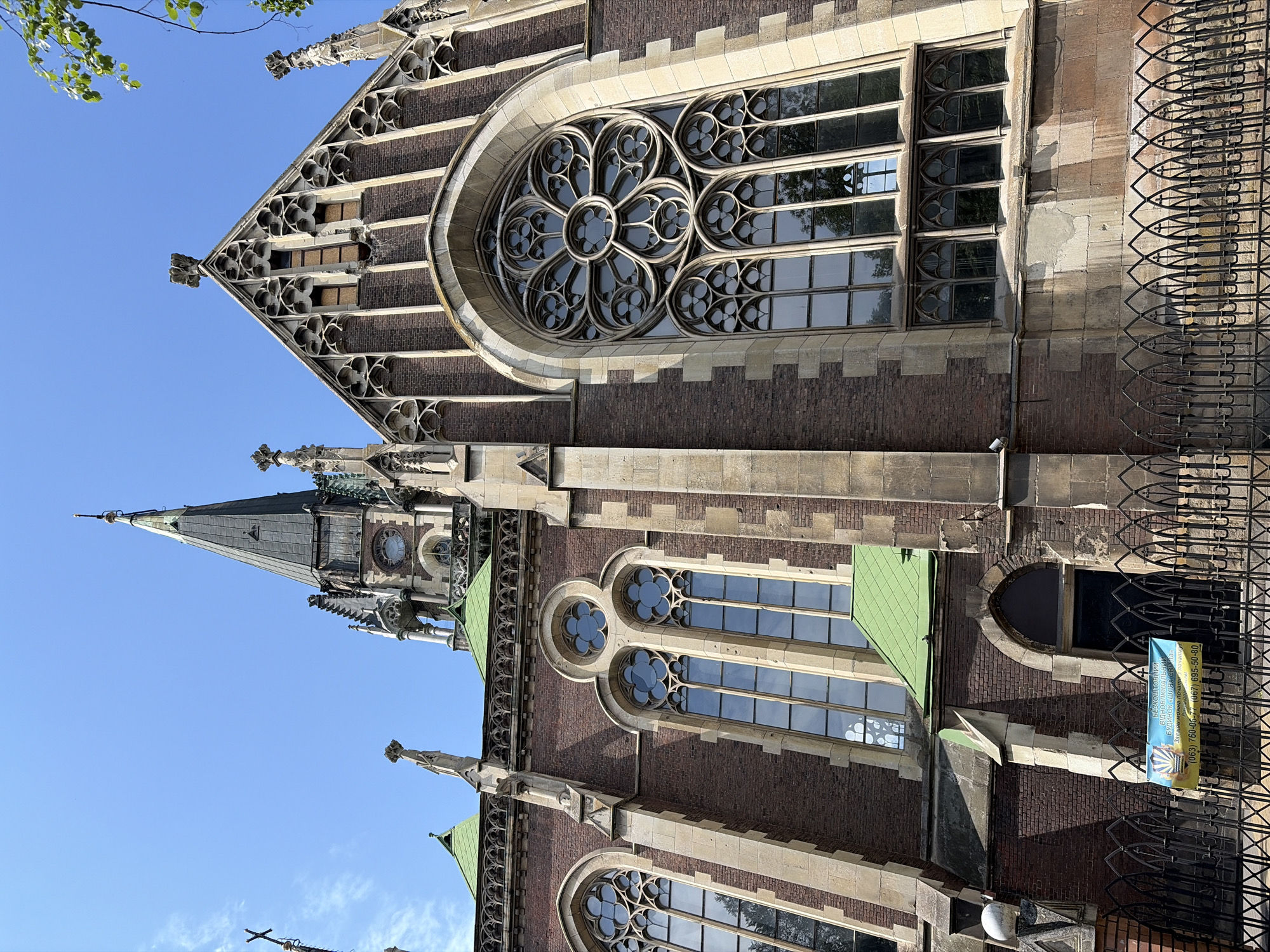 The image shows a low-angle, full shot of the Saint Elizabeth's Church in Lviv, Ukraine, on a clear, sunny day. The church is constructed of dark brick with lighter stone accents, featuring Gothic architectural elements like pointed arches, intricate tracery in the windows, and ornate pinnacles. A tall, dark spire with a cross at its apex dominates the upper portion of the building, partially obscuring a bright blue sky.
The main facade displays a large rose window with geometric patterns, above a series of tall, narrow windows. To the left, a smaller section of the church features more pointed windows and a green-tiled roof. A black metal fence with a Gothic design runs along the bottom of the image, with a blue and yellow banner hanging on it, displaying text in Ukrainian and what appears to be a stylized depiction of wind. The banner has phone numbers printed on it. The ground level is not visible beyond the fence. No people are visible in the frame.
