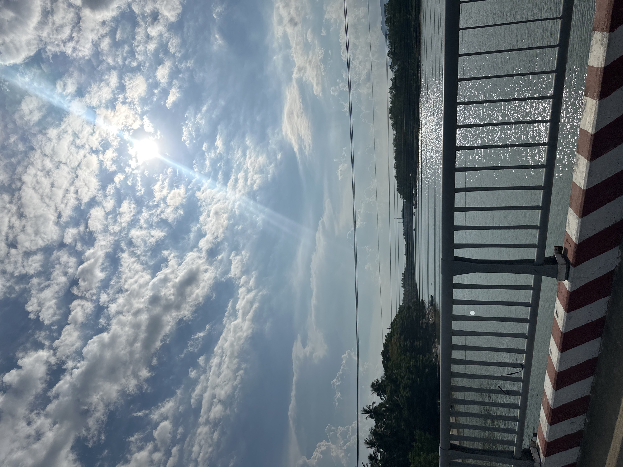 The photograph, taken in Nha Trang Ward, Vietnam, depicts a view from a bridge overlooking a body of water. Sunlight glints off the water's surface. In the foreground, a white and red striped railing is visible. A forested area lines the water's edge. Overhead, the sky is blue with numerous white clouds and a visible sun flare. Power lines are strung across the scene.