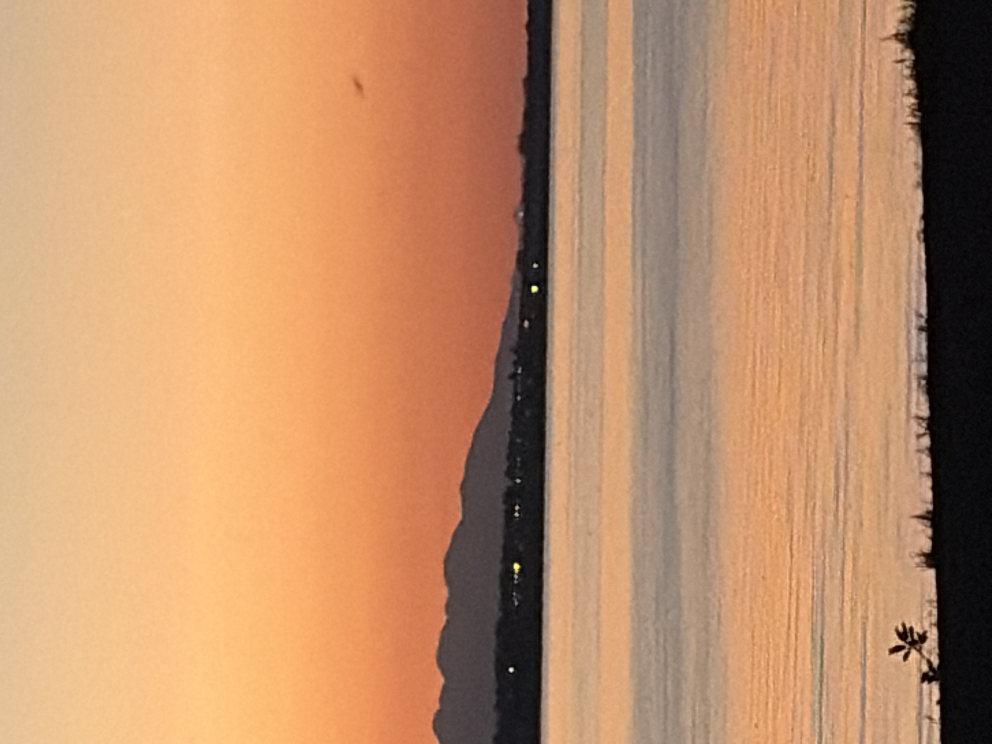 This is a vertical, landscape-oriented photograph capturing a sunset over a body of water. The sky on the left side of the image is a soft gradient of orange and peach hues, suggesting the sun is low on the horizon. A dark, silhouetted mountain range runs along the middle of the frame, separating the sky from the water. Lights, possibly from a distant town or houses, are visible along the edge of the mountains.
The water in the foreground is calm, with distinct horizontal lines created by reflections or subtle ripples. These lines alternate between shades of blue, gray, and orange, reflecting the colors of the sky and the fading light. On the far right of the image, a dark silhouette of what appears to be foliage or branches is visible against the water, adding depth to the scene.
The overall mood is peaceful and serene, characteristic of a sunset. The time of day is clearly evening, during sunset. There are no people or discernible objects in the foreground, other than the natural elements of the landscape. No text is visible in the image. The location is unknown, but the presence of a mountain range and a large body of water could suggest a coastal or lakeside setting.