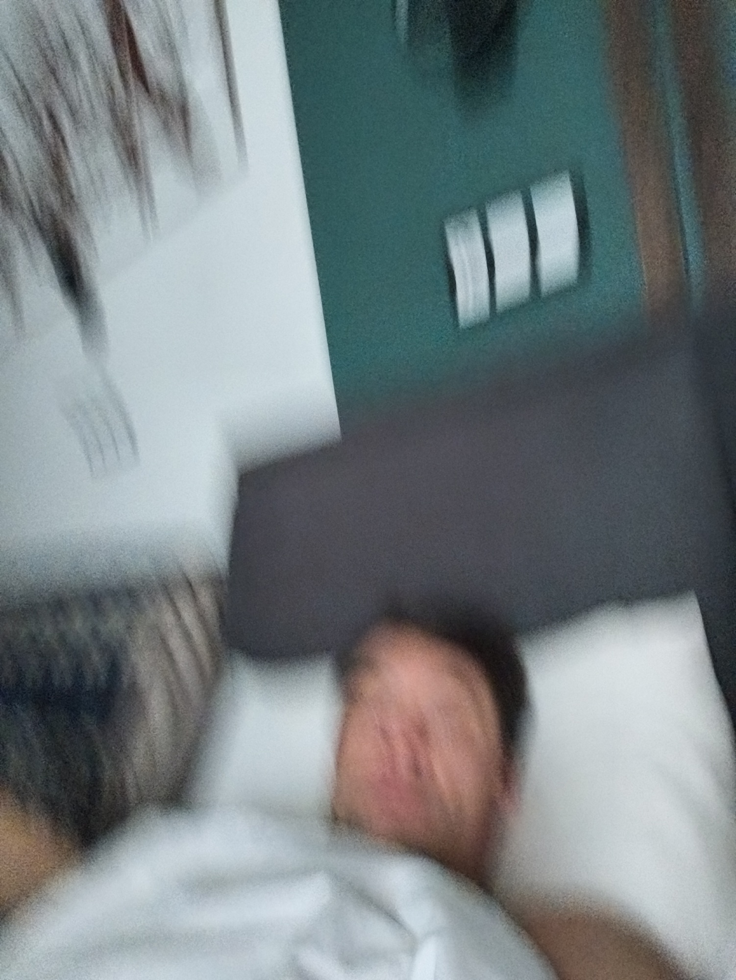 A blurry, low-quality image shows a person lying in bed, presumably indoors. The person is on a white pillow and covered with a white sheet. In the background, there is a teal wall with three light switches or outlets. To the left is a light-colored wall with abstract decor. Due to the blur and lack of detail, it's impossible to determine the location or any specific actions taking place. The mention of Moscow, Russia is not evident in the image.