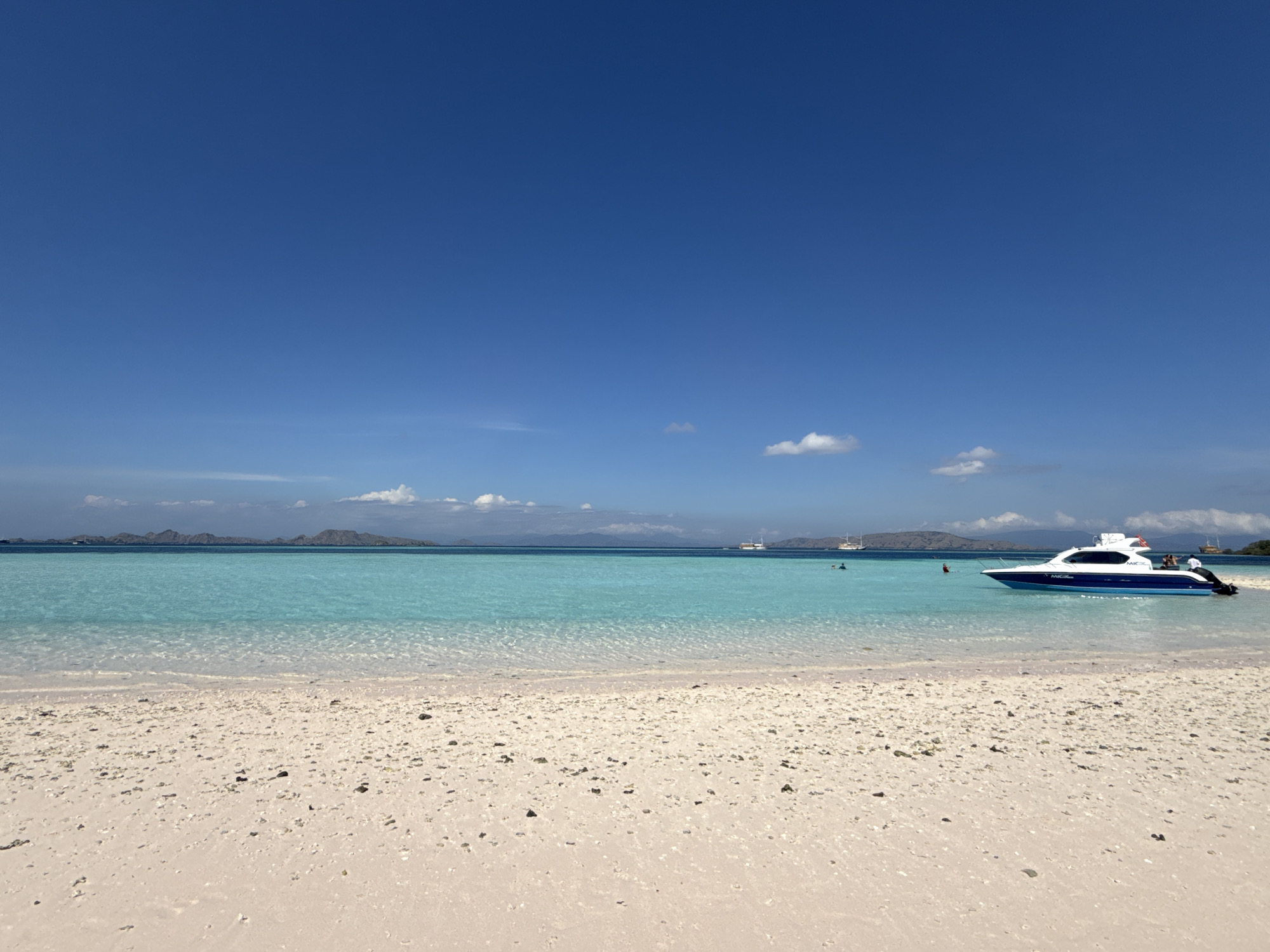The image depicts a beach scene in Komodo, Indonesia, under a clear blue sky with sparse clouds. The beach has light-colored sand speckled with small rocks and shell fragments. The turquoise water is clear near the shoreline, with visible undulations. In the middle distance, there are small, hilly islands. A white motorboat with blue accents is anchored in the shallow water. Several distant vessels are visible. There are people swimming in the water.