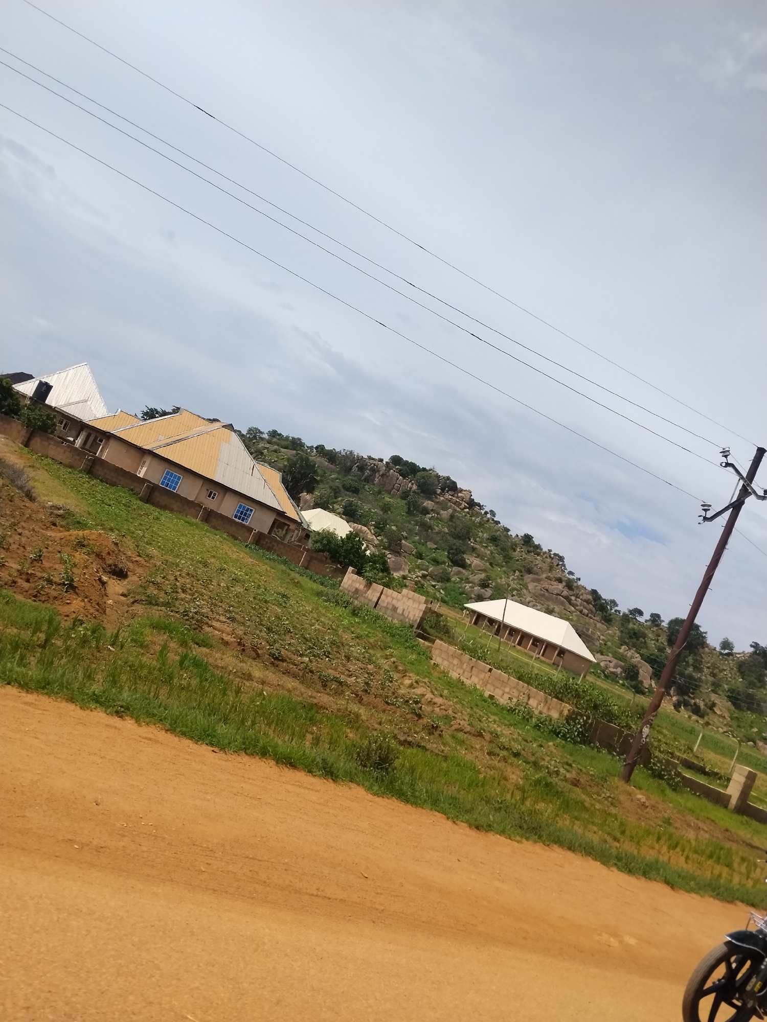 The image depicts a rural hillside scene in Nabatum, Nigeria, under an overcast sky. In the foreground, a dirt road with a reddish-brown surface is visible, with a portion of a black motorcycle's wheel in the lower right corner. The mid-ground features a terraced hillside with patches of green grass and cultivated soil. Several buildings are situated on the hillside. One prominent building, to the left, has a light-colored, multi-gabled roof and light-colored walls with blue-framed windows. Another building, further to the right and lower on the hill, has a white hip roof and a colonnaded walkway. A brick wall runs along the edge of the property containing these buildings. In the background, a rocky, tree-covered hill rises steeply, with more buildings scattered on its slopes. A power line with insulators runs diagonally across the sky, supported by a wooden pole on the right side of the image. The overall impression is one of a developing rural community in a hilly landscape.
