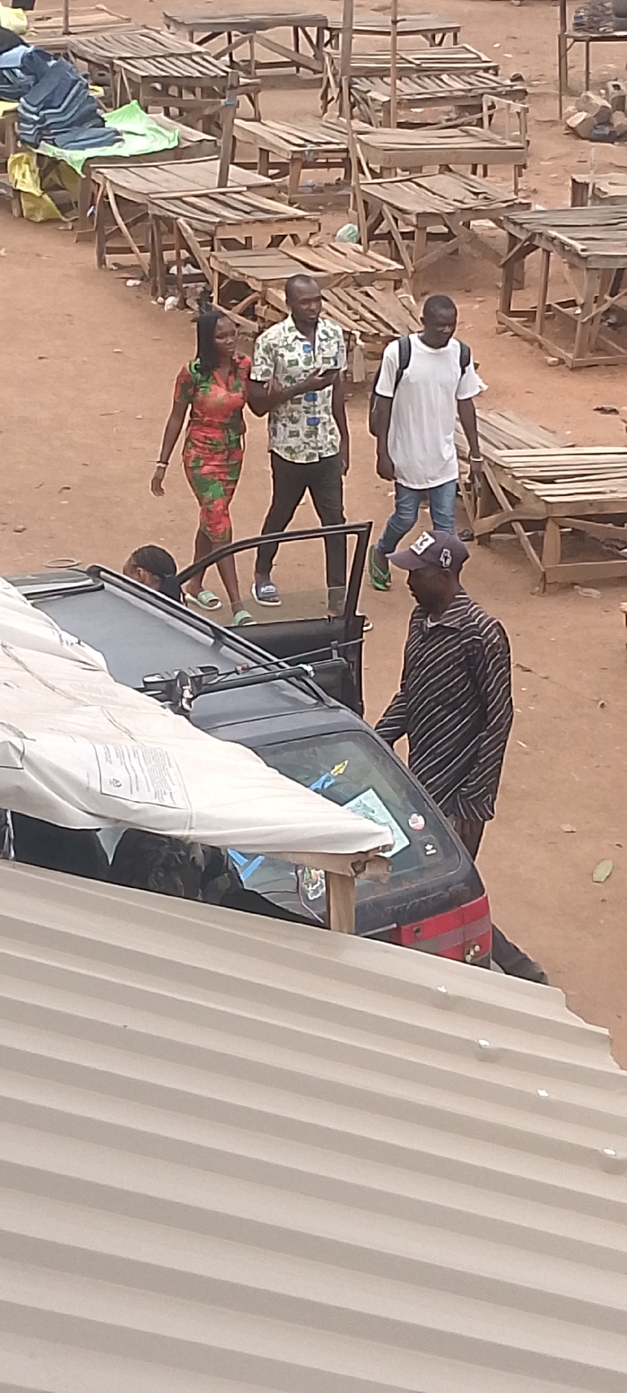 The image depicts an outdoor scene in Bukuru, Nigeria. A black car with an open driver-side door is visible, with a man in a striped shirt and cap standing next to it. Another person is partially visible inside the car. In the background, a woman in a floral dress is walking arm-in-arm with a man in a patterned shirt. A third man in a white t-shirt and jeans is walking behind them. Numerous empty wooden tables are arranged in rows in the background, suggesting a marketplace setting. The ground is reddish-brown, and part of a corrugated metal roof is visible in the foreground.