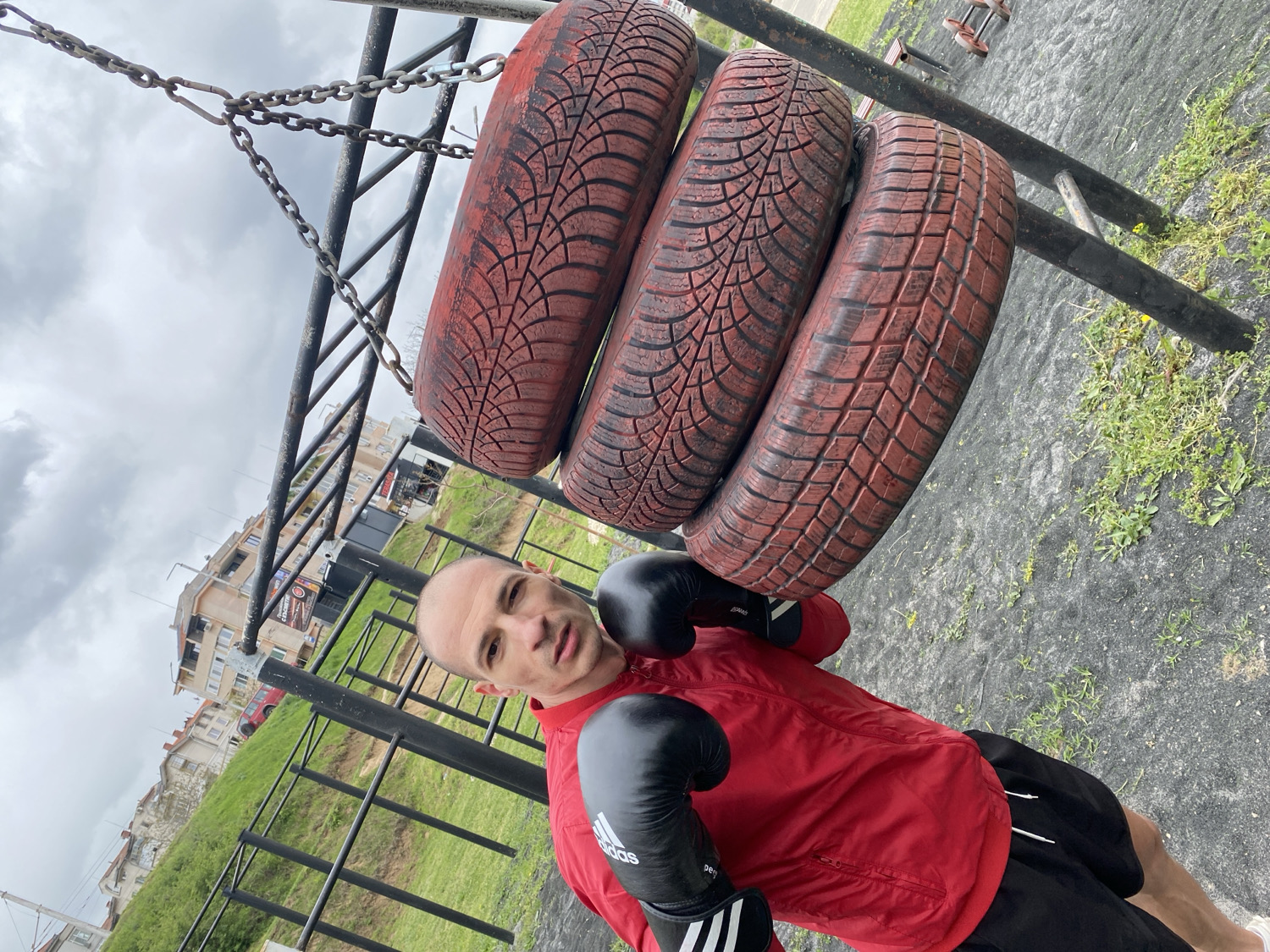 An outdoor, portrait-oriented shot captures a man posing in what appears to be a training area in Pleven, Bulgaria. The man, with a shaved head and light skin, looks directly at the camera with a neutral, focused expression. He is wearing a red long-sleeved athletic jacket, black shorts, and black boxing gloves, one of which clearly displays the Adidas logo.
To his right, three reddish-brown car tires are stacked vertically and suspended by metal chains from a black metal framework, resembling a pull-up bar or an improvised gym structure. The tires feature distinct tread patterns, indicative of winter tires. The man's attire, particularly the boxing gloves, suggests he is engaged in or preparing for a boxing or striking workout session, possibly using the suspended tires as a punching bag alternative.
The ground beneath the structure is covered with a dark, textured material, possibly rubber matting or geotextile fabric, with patches of green grass and weeds growing through it. In the mid-ground, a grassy slope leads up to a cluster of multi-story buildings, which appear to be residential with visible balconies, setting the scene in an urban or suburban context. The sky is overcast and cloudy, indicating daytime on a dull day. A blurry sign with text is partially visible on one of the distant buildings.