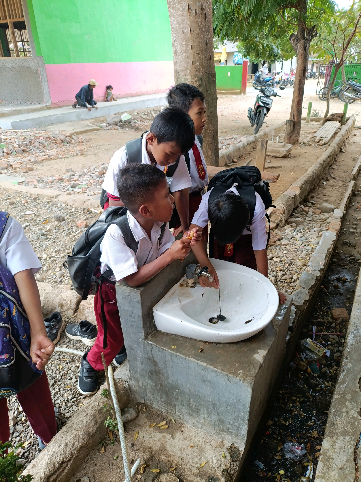 In an outdoor area of a school in Sumberejo, Indonesia, four young students wearing school uniforms are gathered around a white sink. The sink is mounted on a concrete pedestal, which is next to a dirty, open drain filled with debris and water. Two of the students, a boy and a girl, are standing. The boy in the foreground is wearing a white shirt, maroon pants, and a black backpack. He is leaning over the sink, holding a piece of orange in his right hand and a thin string with two dark objects attached to it in his left hand, which he is dangling into the sink. The girl, partially visible on the far left, wears a blue backpack with a pattern and maroon pants.
Behind them, two more boys are standing. The boy to the left of the first boy is wearing a white shirt, maroon pants, and a black backpack. He is looking down into the sink. The boy further back, also in a white shirt and maroon pants, is mostly out of focus. In the background, a green and pink building is visible, along with a tree with lush green leaves. Several motorcycles are parked in front of another building further in the distance. The ground is covered with dirt, gravel, and scattered debris.