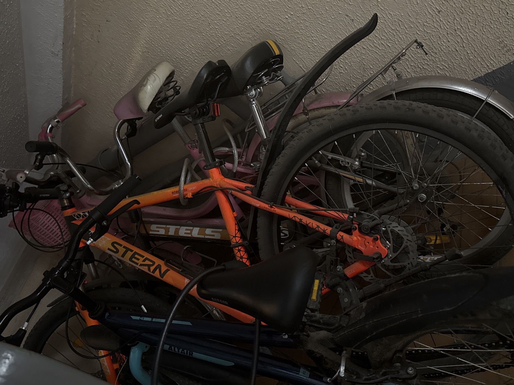 A cluttered image shows a collection of several bicycles stored indoors. The bicycles are various models and colors, including pink, orange, and blue. One of the bikes is labeled "STELS" and another "STERN." A third is labelled "ALTAIR". The setting appears to be a storage area, possibly a garage or basement, with a textured wall visible in the background. The location is likely Mytishchi, Russia, due to the presence of the Russian bicycle brand "STELS."