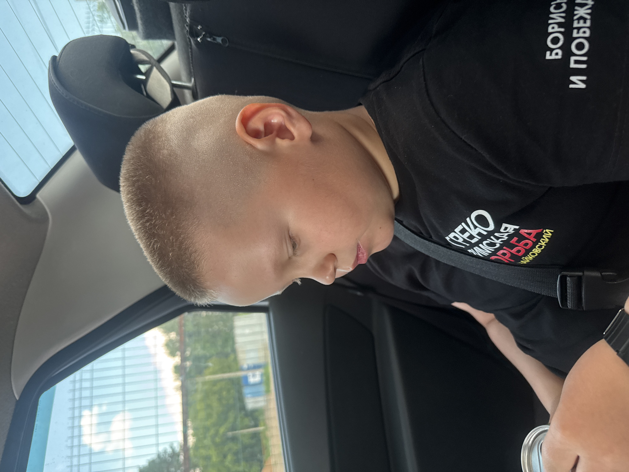 A young boy with a buzz cut is seated in the back of a car. He is wearing a black t-shirt with white and red lettering. He is looking down at something in his lap. A can and a watch are visible on his wrist. The interior of the car is dark, with a black seat visible. Through the window, a fence and some trees can be seen, suggesting an outdoor setting. The scene appears to be in Chaikovsky, Russia.